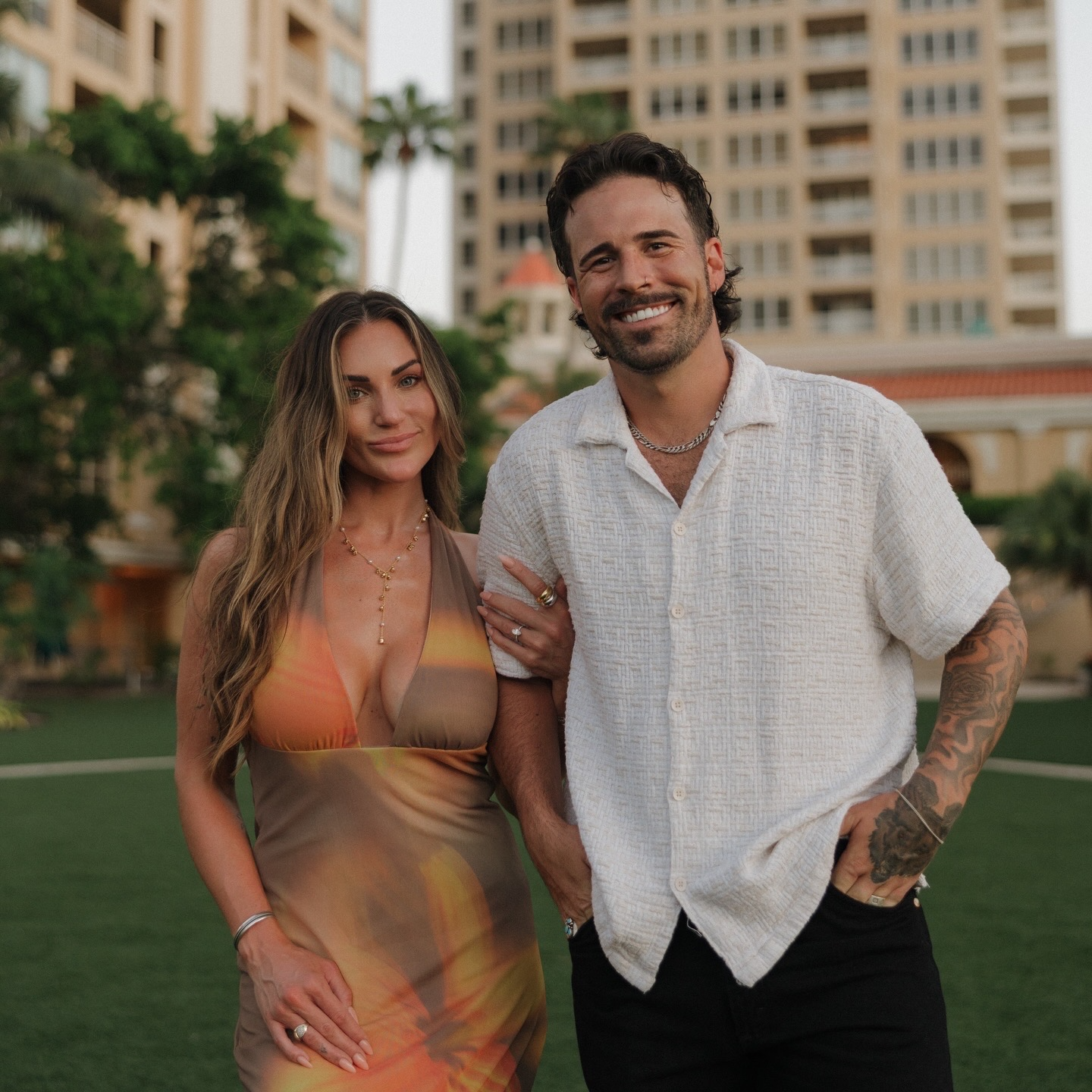 A smiling couple standing outdoors on a grassy area with tall buildings and palm trees in the background during sunset.