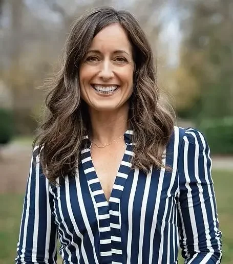 A woman with long brown hair, wearing a blue and white striped blazer, smiling outdoors with blurred trees in the background.