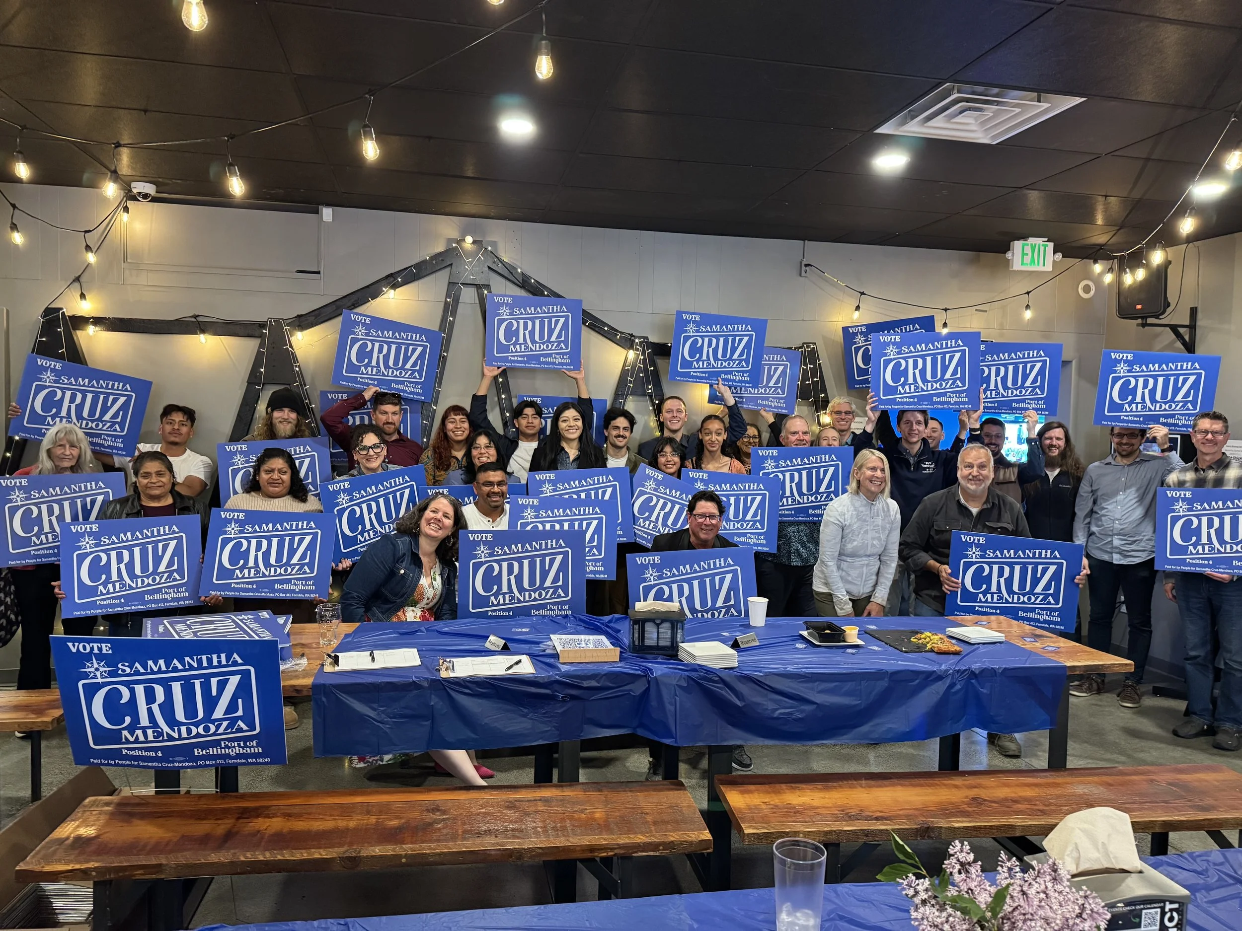 Group of people holding blue campaign signs that say 'Vote Samantha Cruz Mendoza, Port of Bellingham,' in an indoor event with hanging lights, tables, and decorations.
