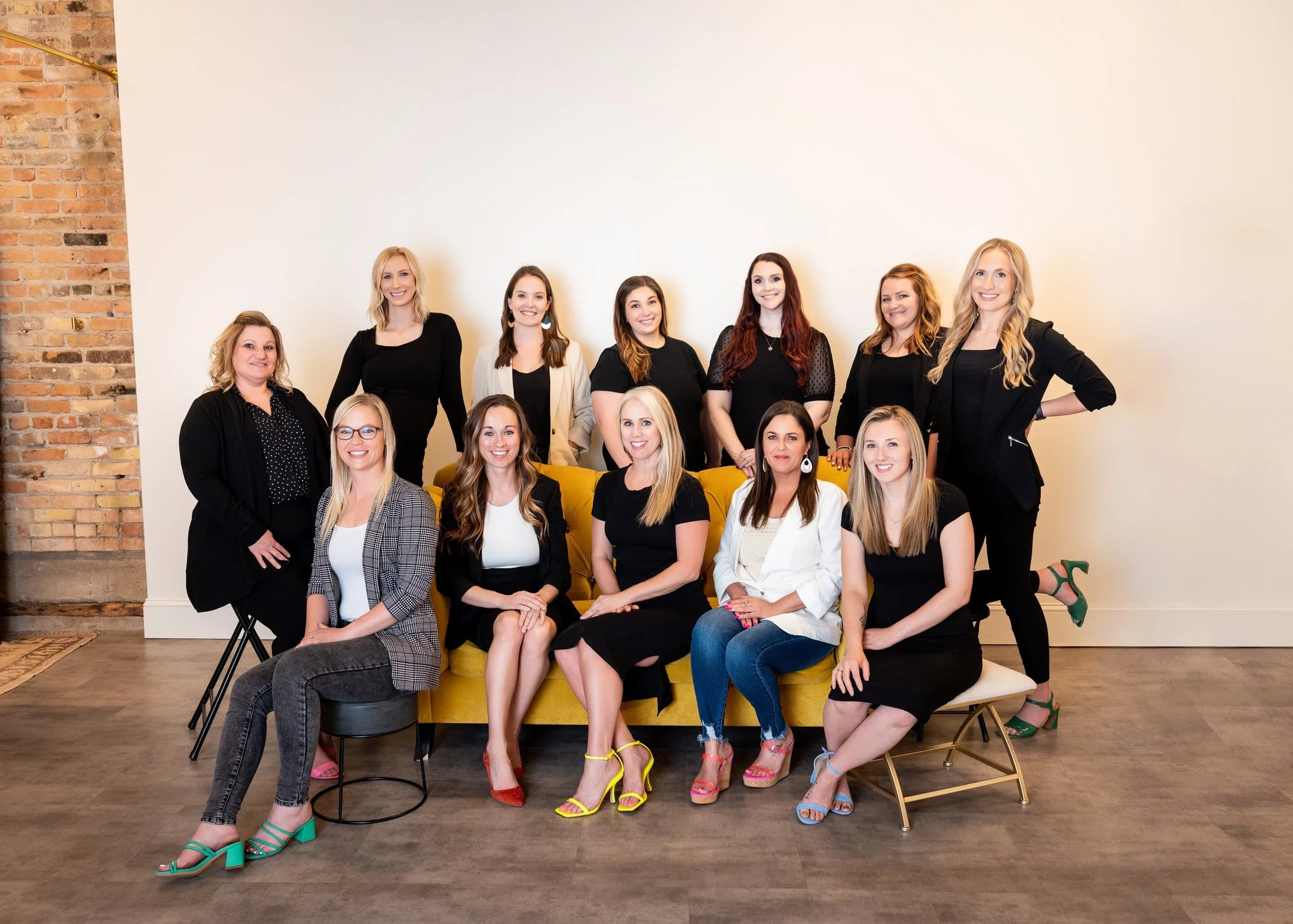 A group of eleven women posing together in a studio setting with a plain wall and brick wall in the background. Some are standing, some sitting on a yellow sofa, and some are seated on various stools and chairs. They are dressed in a mix of black, white, plaid, and beige clothing, with some wearing colorful high heels. Everyone is smiling and looking at the camera.