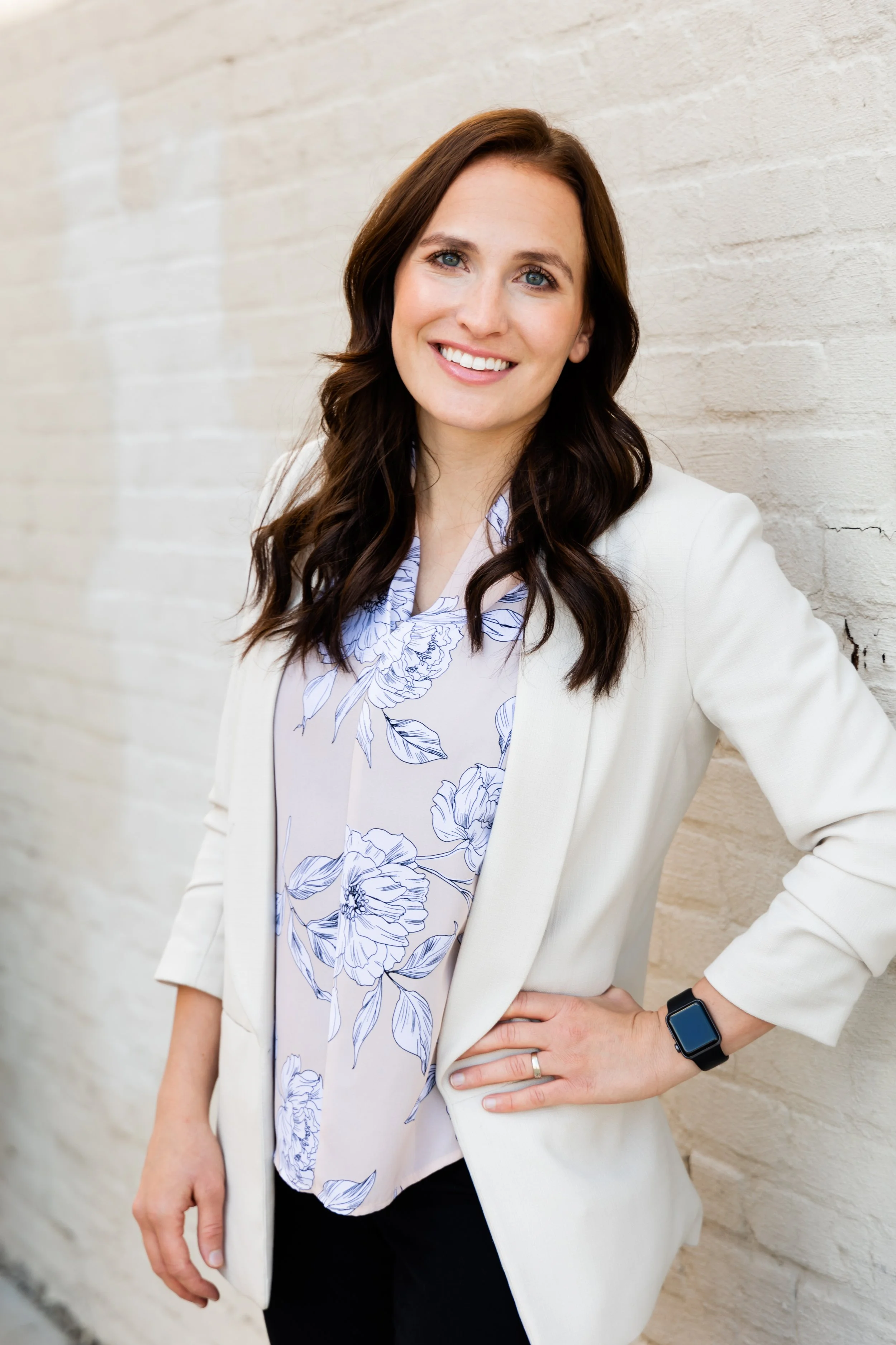 A woman with long brown hair, blue eyes, and a bright smile, standing against a white brick wall, wearing a beige blazer, a floral blouse, a smart watch, and a ring on her left hand.
