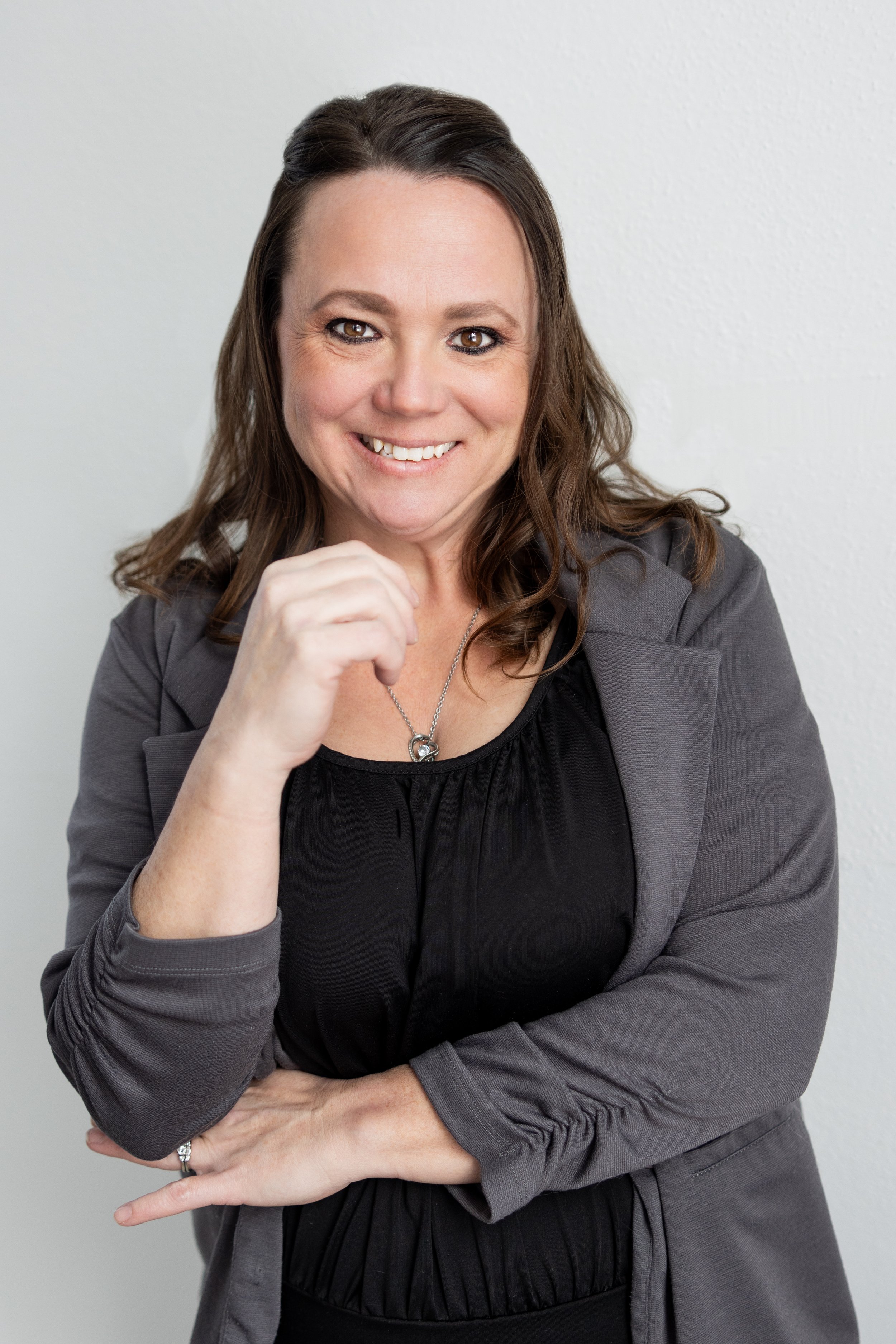 A woman with brown hair, smiling, wearing a gray blazer over a black top, with a silver necklace and rings, standing with her arms crossed against a white background.