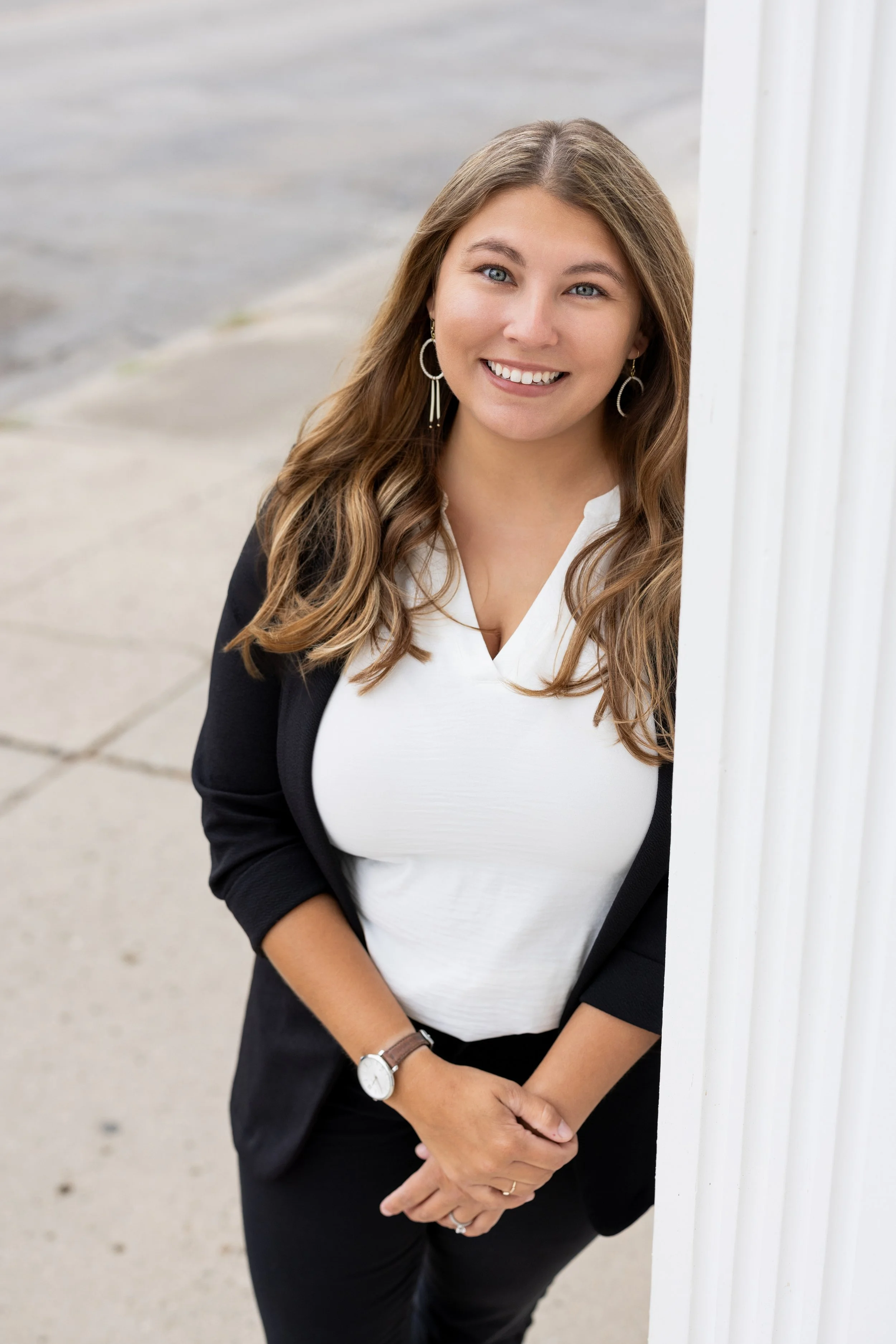 Portrait of a smiling woman with long brown hair, wearing a black blazer, white shirt, earrings, and wristwatch, standing outdoors next to a white column.