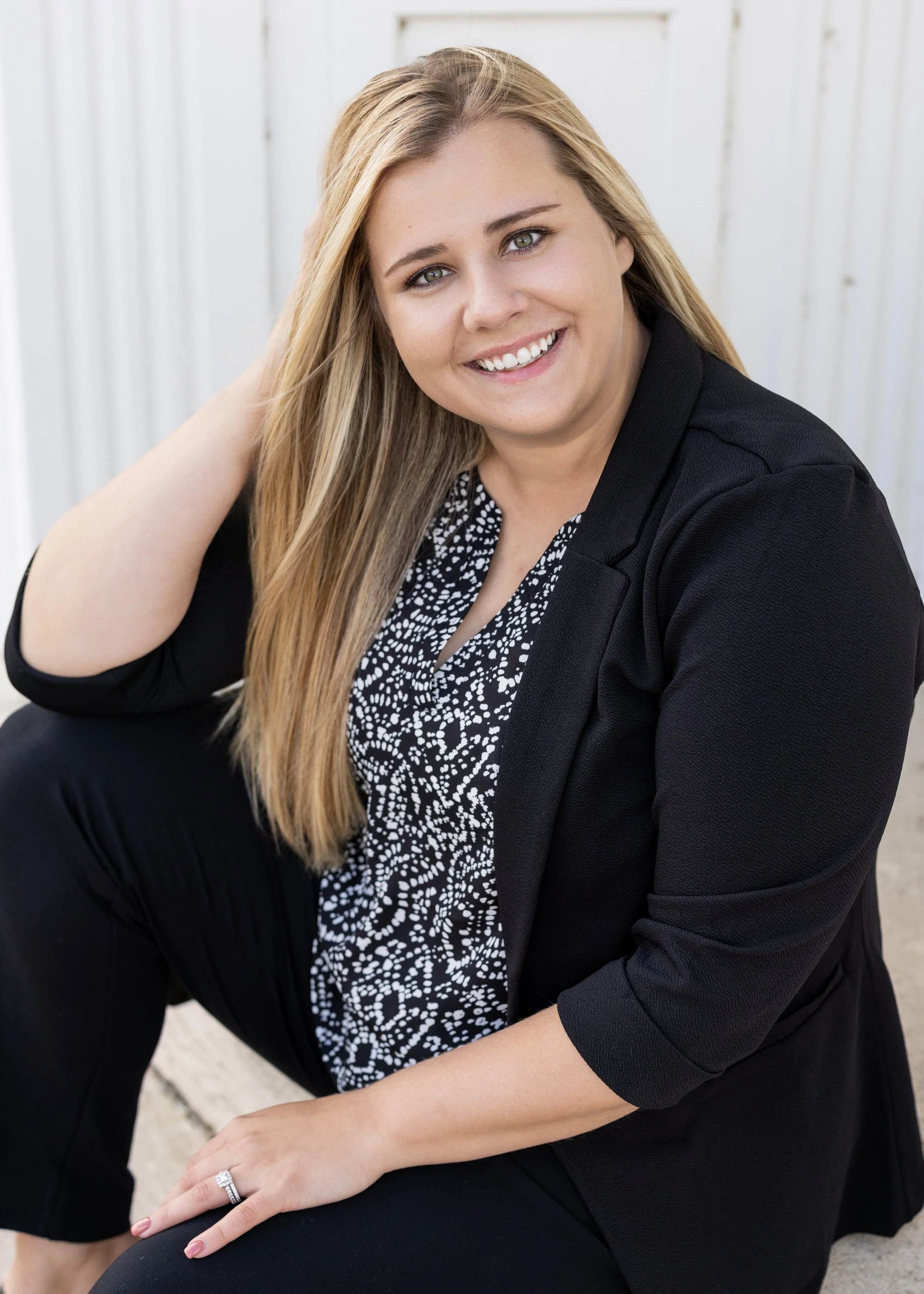 A smiling woman with long blonde hair, wearing a black blazer over a black and white patterned blouse, sitting outdoors in front of a white wooden fence.