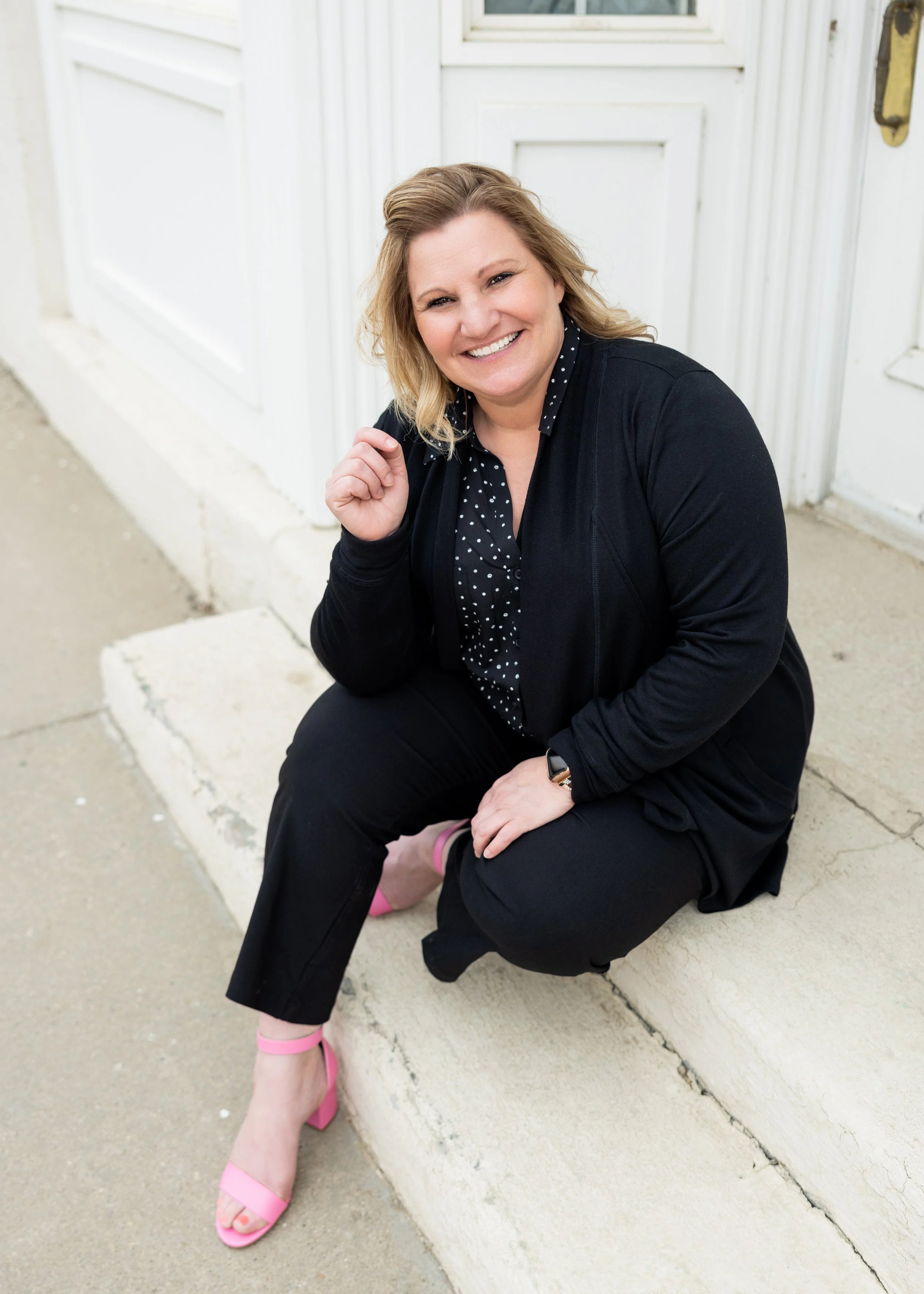 A woman with blonde hair smiling, sitting on the ground with one knee up, wearing a black jacket, black pants, a polka dot shirt, pink high heels, and a smartwatch, in front of a white building with a white door and window.