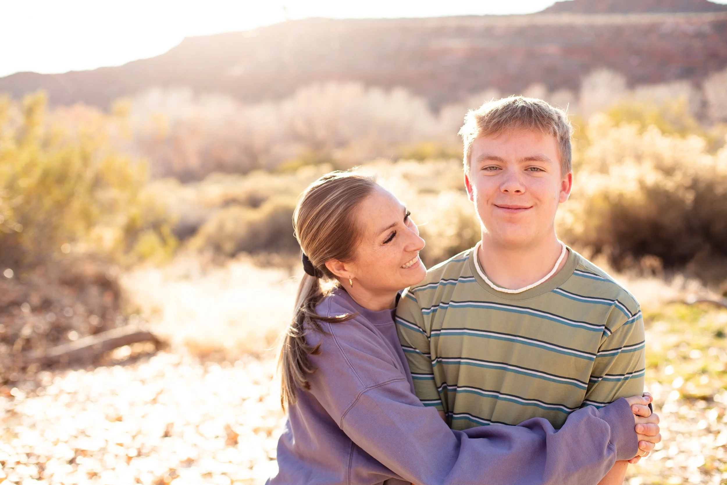 A woman and a teenage boy embracing outdoors in autumn sunlight, with mountains and yellow leaves in the background.