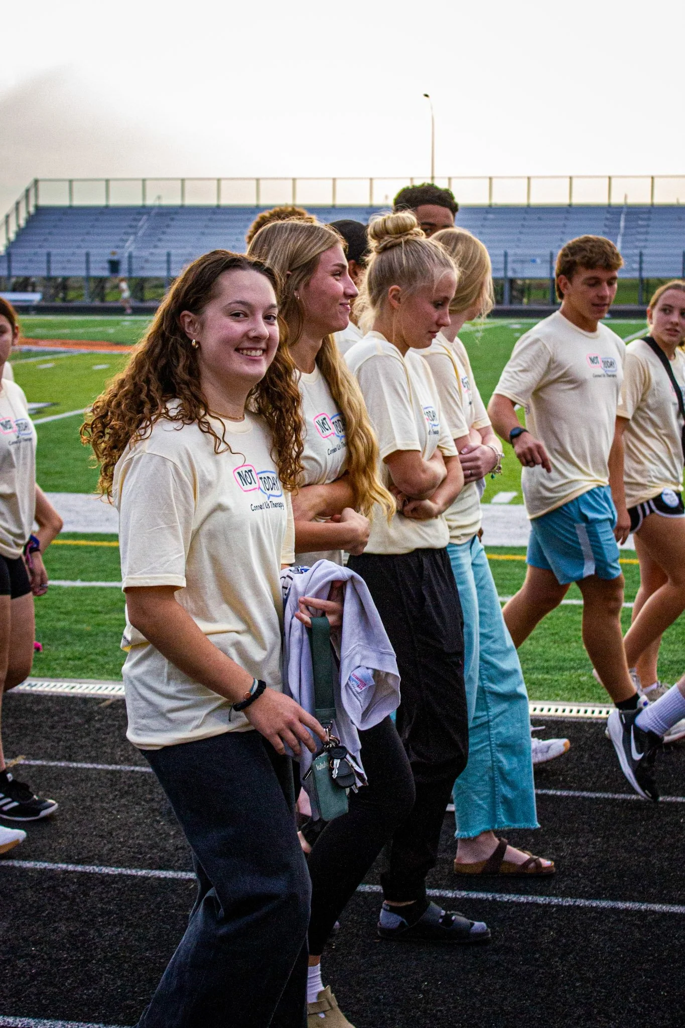 A group of young people walking on a track field during sunset, wearing matching beige T-shirts with text and graphics, some smiling and some looking ahead.