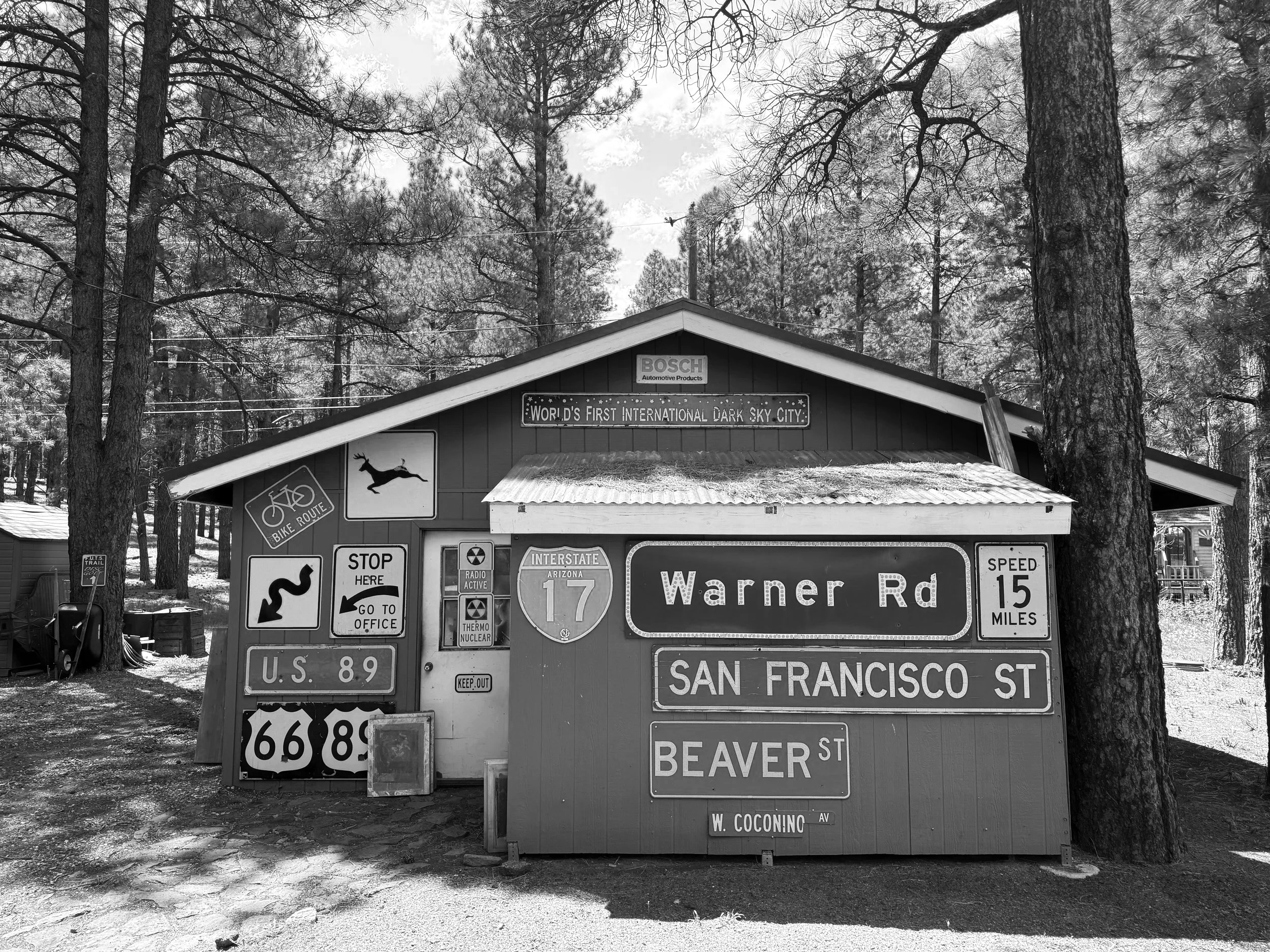 A building with a sign indicating it is the 'World's First International Dark Sky City,' surrounded by trees. The building has various road and traffic signs, including route numbers and directions, with a wooded background.