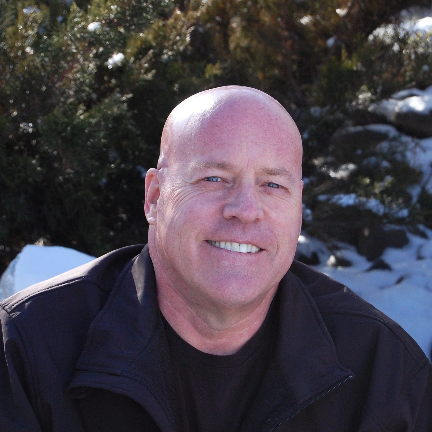 A smiling, bald man with light-colored eyes, wearing a black shirt and black jacket, outdoors with snow and trees in the background.
