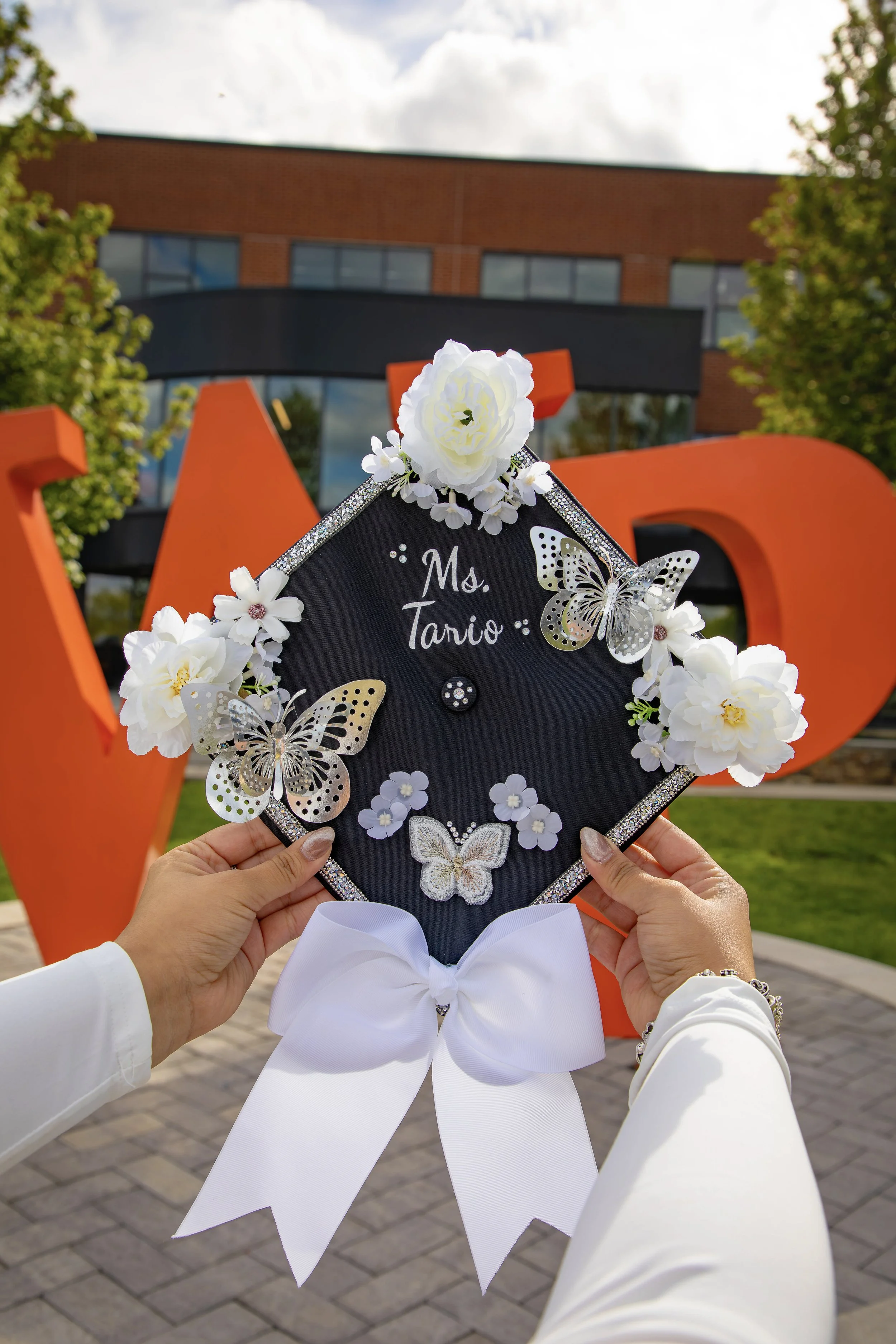 A person holding a decorated black graduation cap with white flowers and butterflies, reading 'Ms. Tavio'. In the background, large orange letters spell 'LOVE' against a building with trees and cloudy sky.
