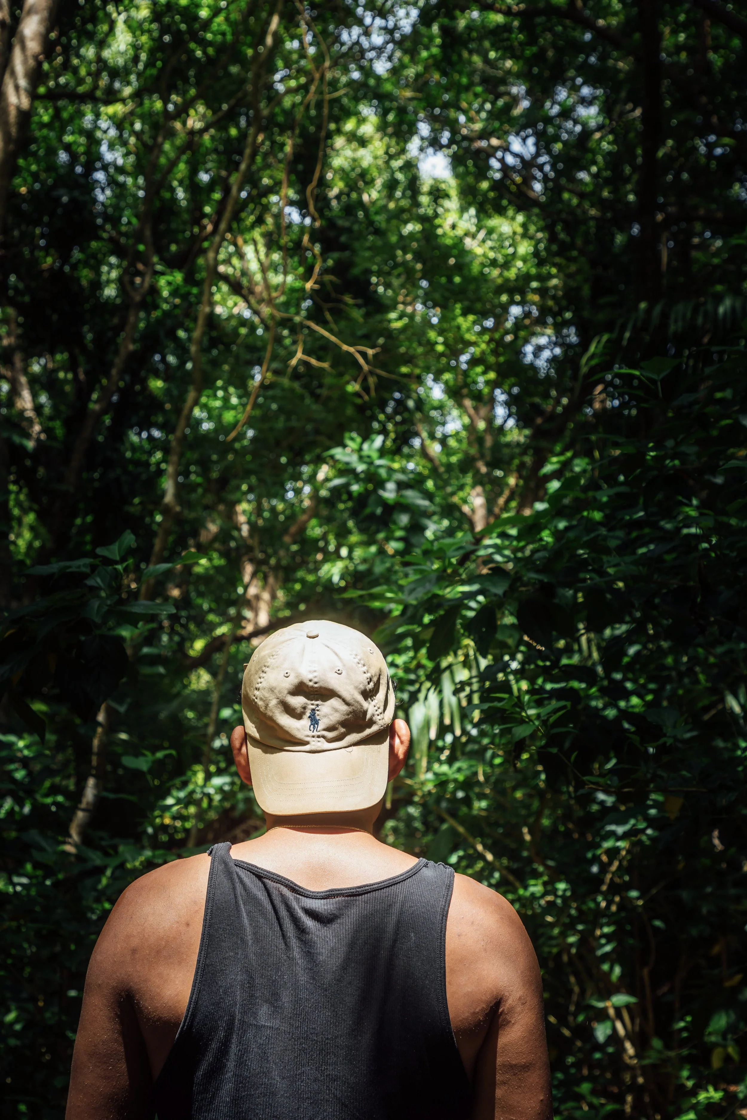 A person with dark skin wearing a beige cap and a black tank top, seen from the back, standing in a lush green forest canopy with sunlight filtering through the leaves.