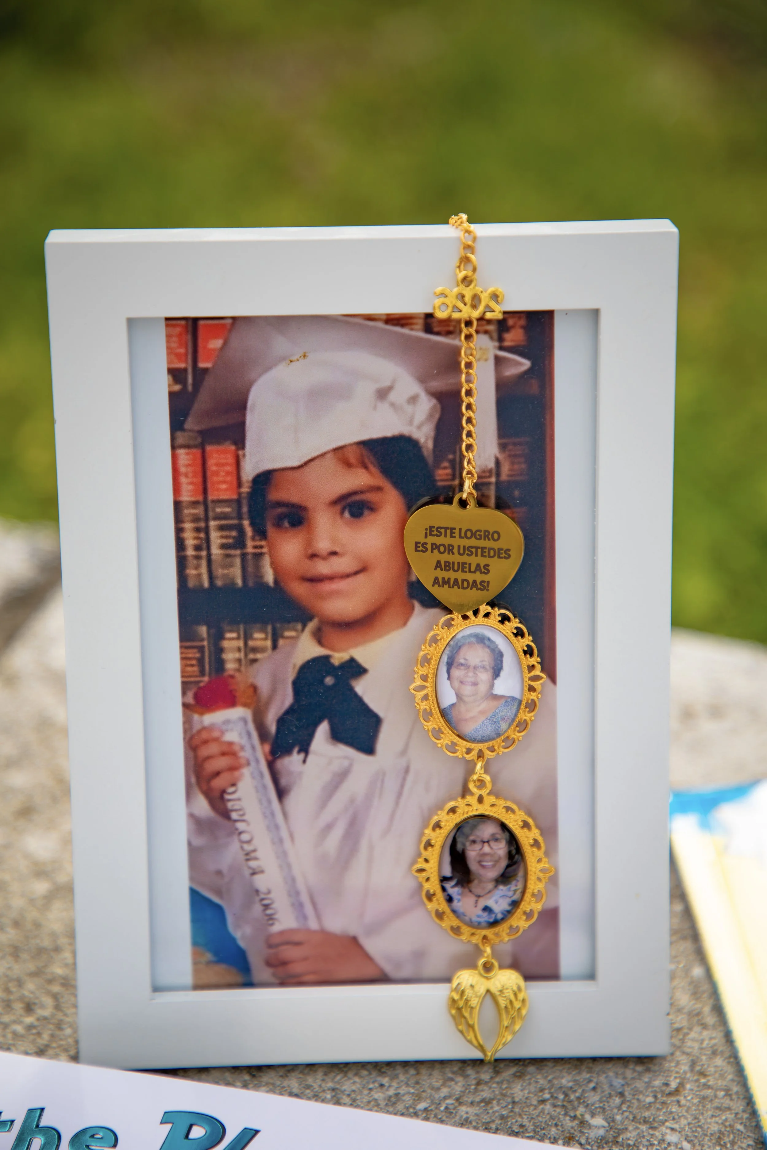 A gallery photo shows a framed picture of a young girl in a graduation cap and gown, holding a diploma, with a bookcase in the background. Hanging from the top right of the frame is a gold rosary with a heart-shaped badge that has a message in Spanis
