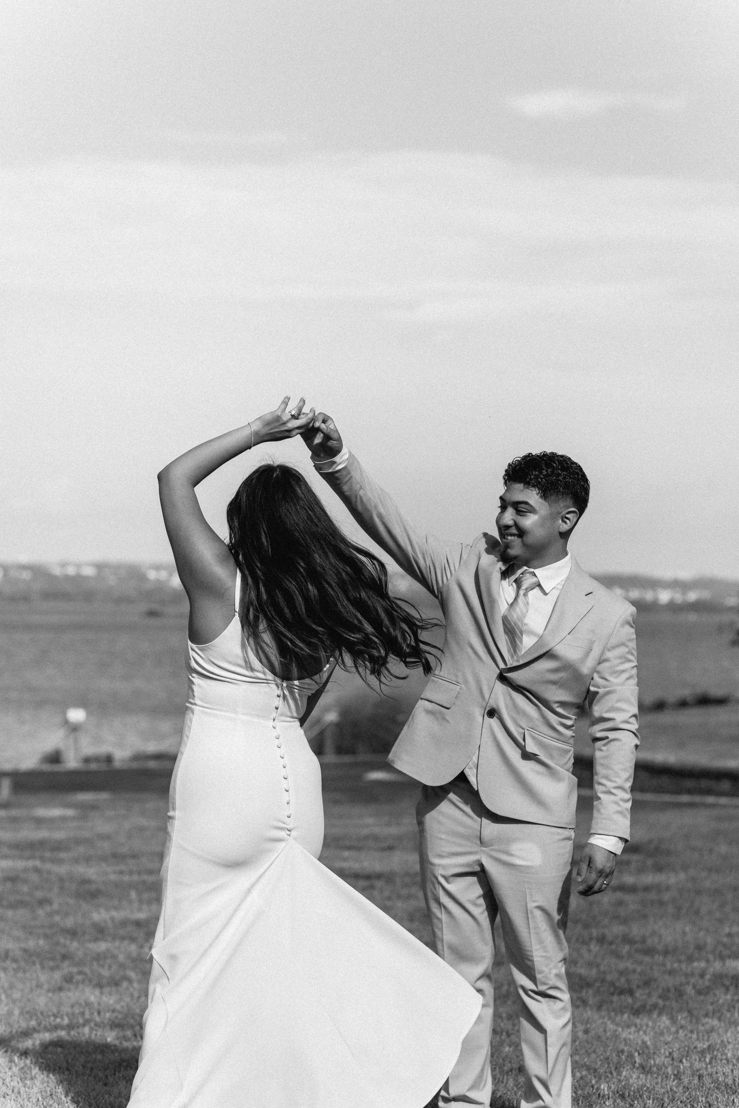 A couple dancing outdoors on a grassy field, with the man in a light-colored suit and the woman in a long dress. They are holding hands and smiling, with open land and cloudy sky in the background.