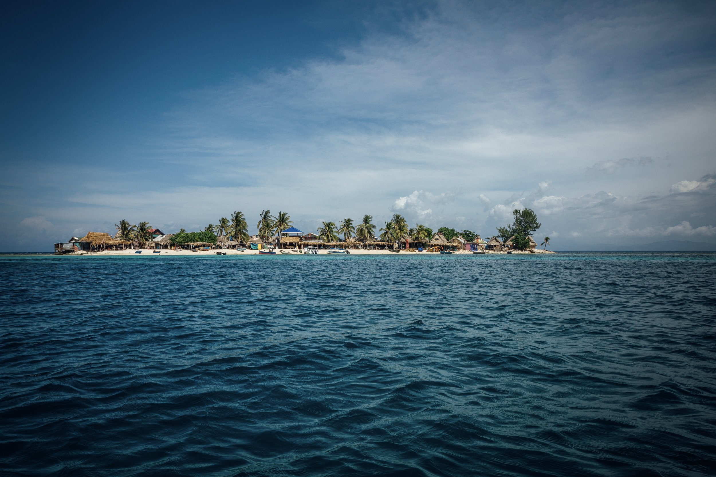 A tropical island with a sandy beach, palm trees, and thatched-roof huts, viewed from the ocean with clear blue water.