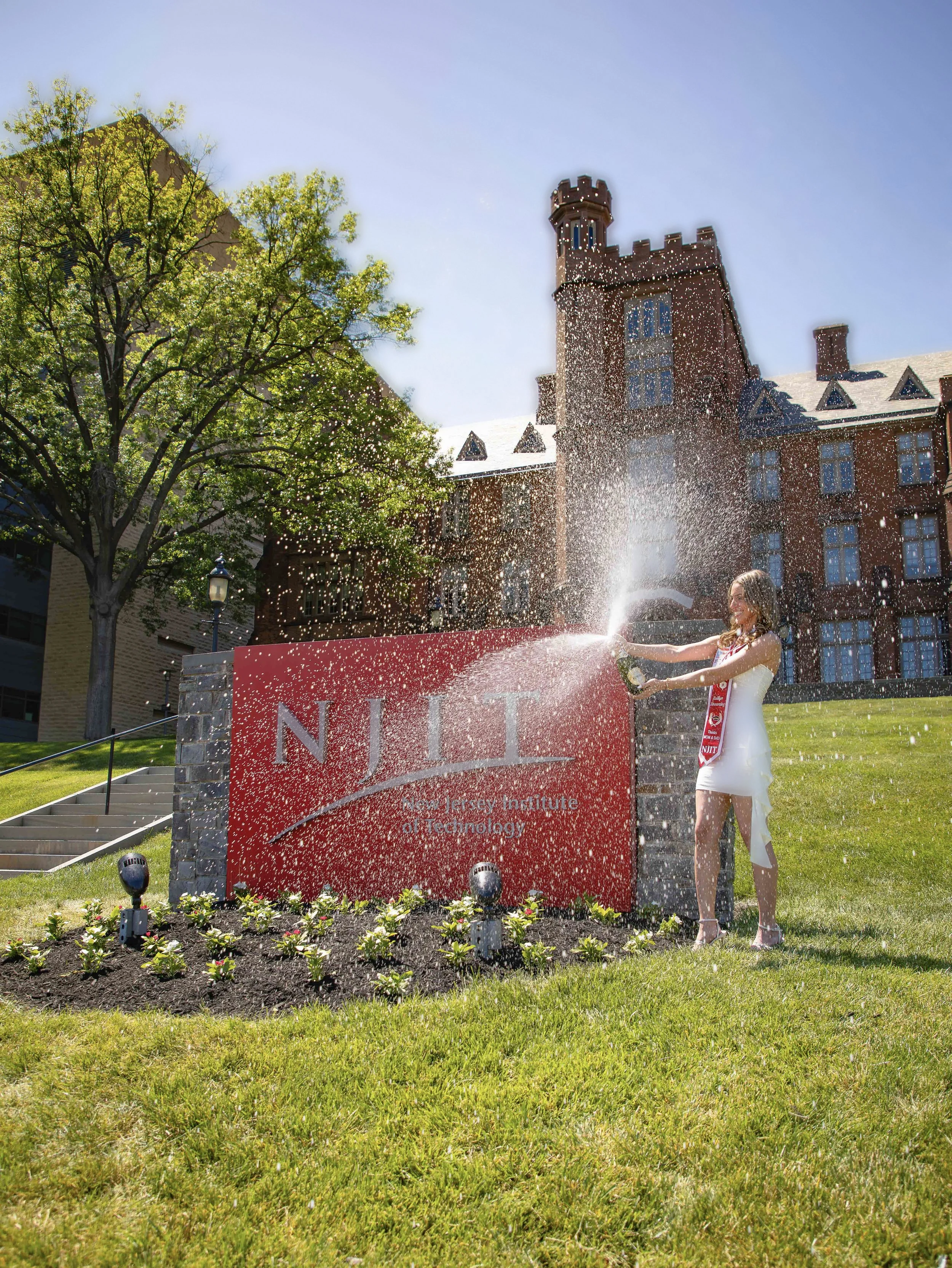 Students celebrating graduation at the New Jersey Institute of Technology, with a fountain, green lawn, and historic brick building in the background.