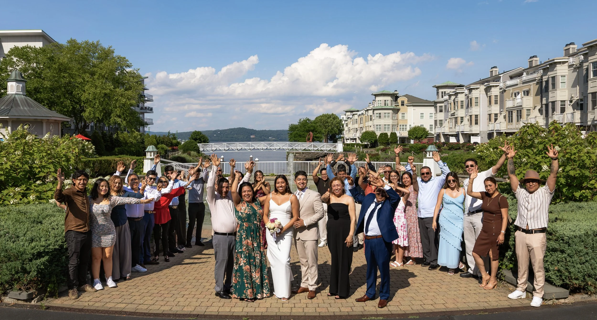 Group of people gathered outdoors on a sunny day near a waterway with greenery, buildings, and hills in the background, celebrating and smiling.