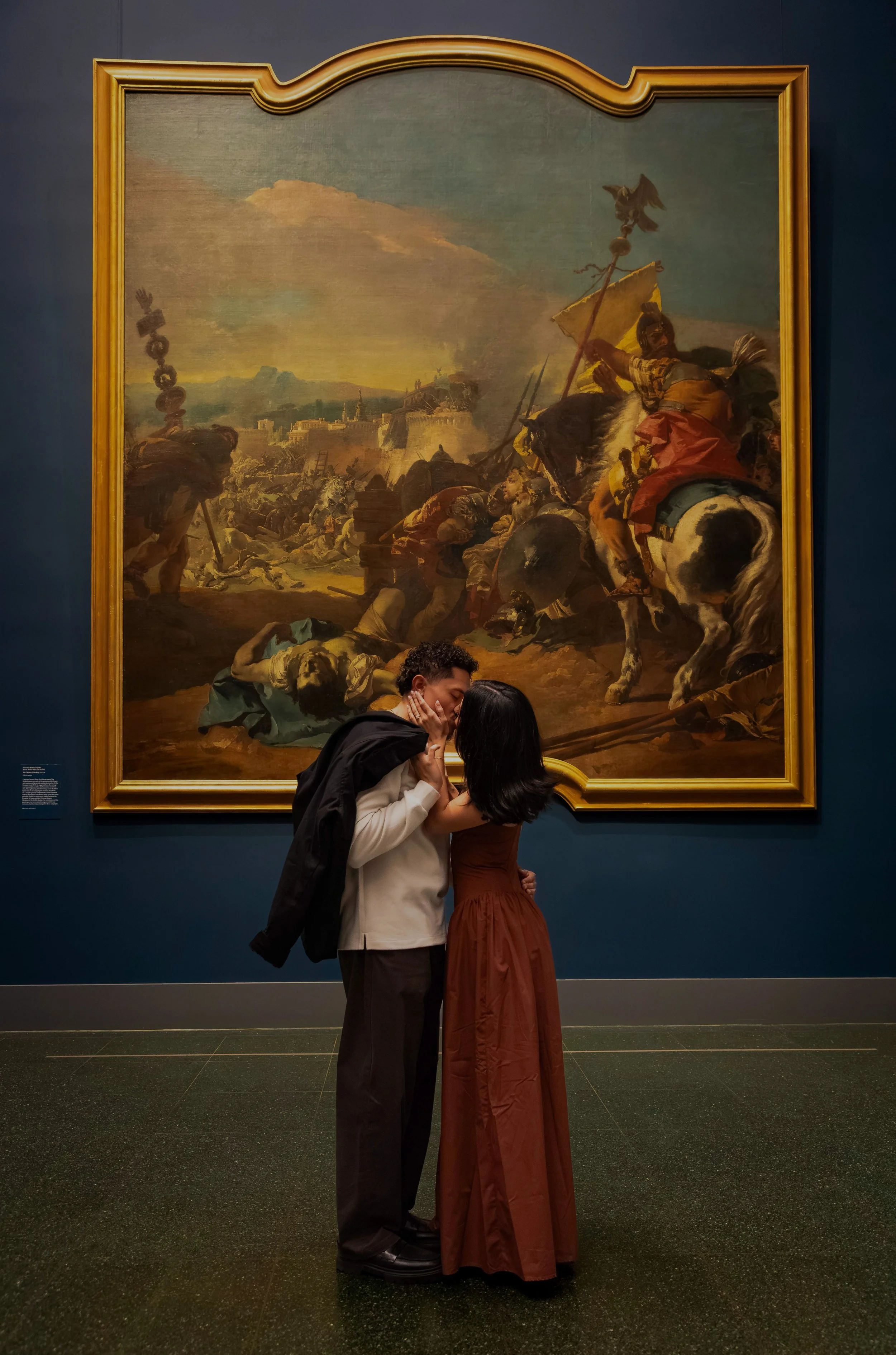 Two people hugging in front of a large painting in an art gallery. The painting depicts a historical battle scene with soldiers, horses, and a cloudy sky.