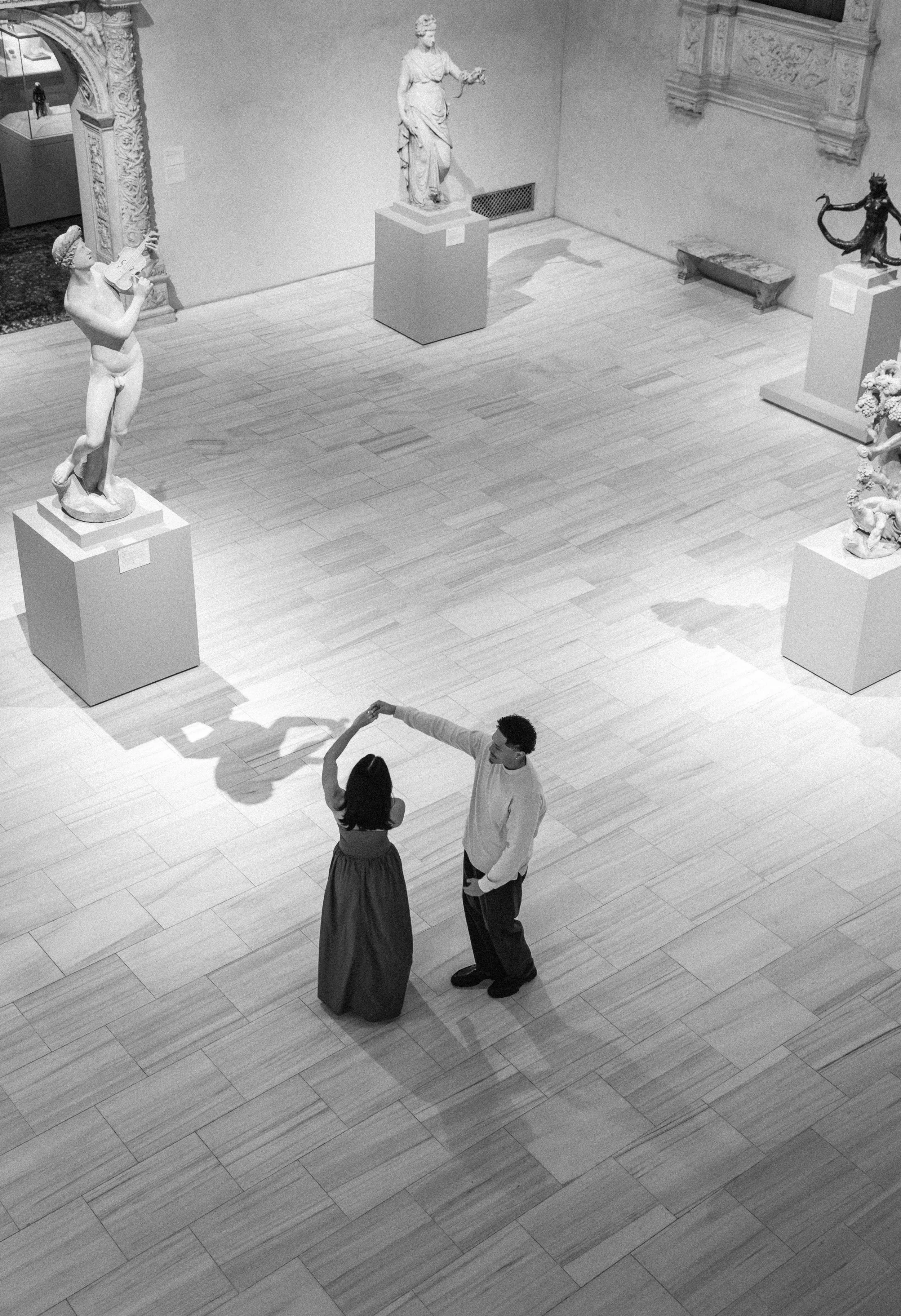 Two couples dancing in an art museum gallery with classical sculptures on display, including a statue of a woman with a helmet and a nude male sculpture playing a musical instrument.