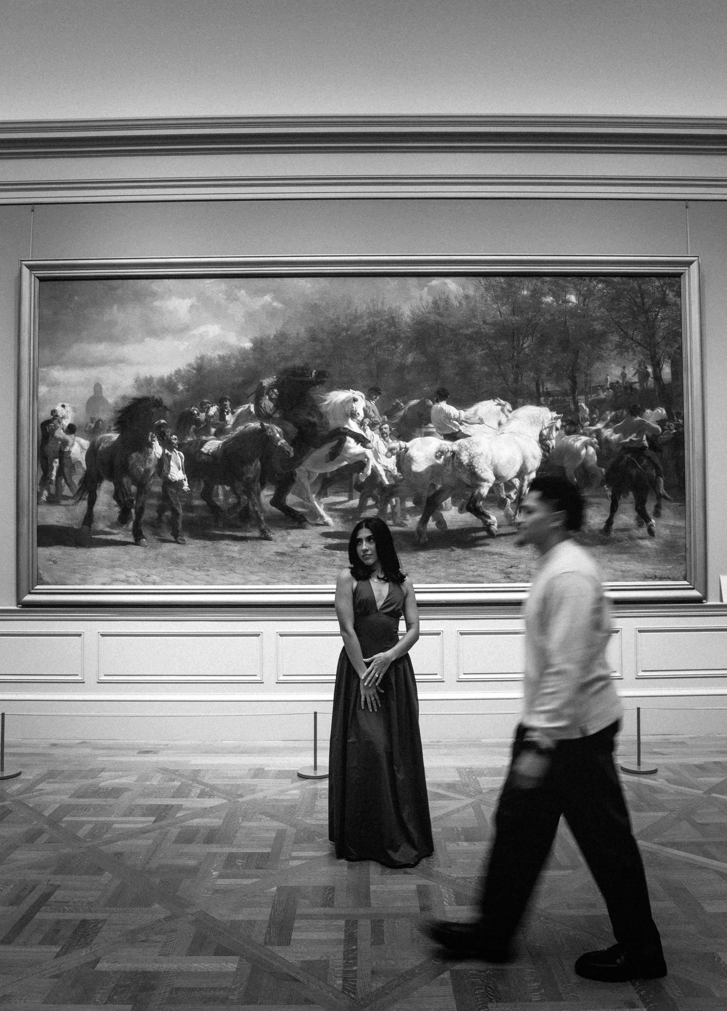 A woman in a long dress stands in front of a large painting of a horse race at an art gallery, with blurred people walking in the foreground.