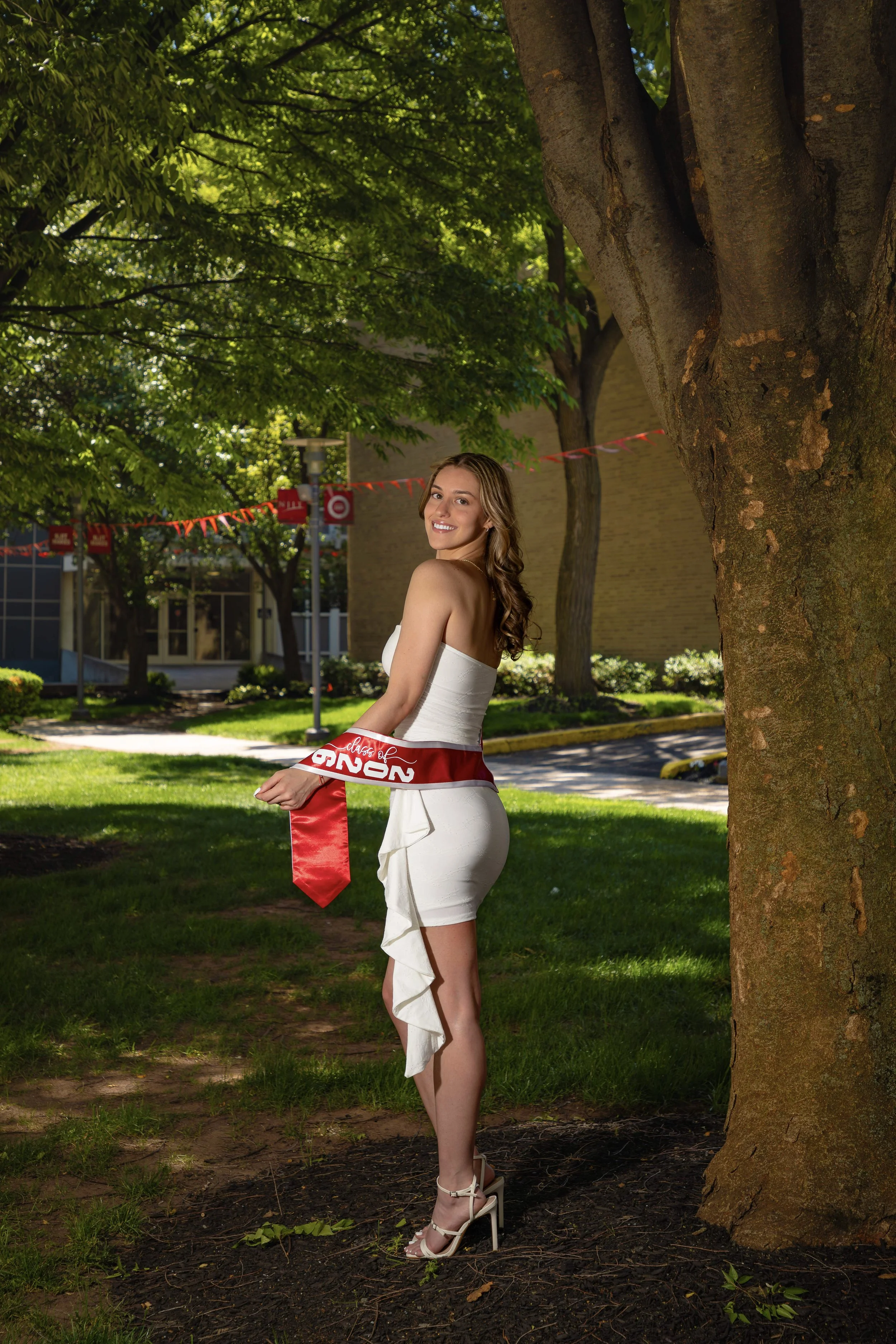 A woman in a white dress and high heels standing outdoors next to a large tree, holding a red sash that reads "Class of 2023" on a sunny day with green trees and a building in the background.