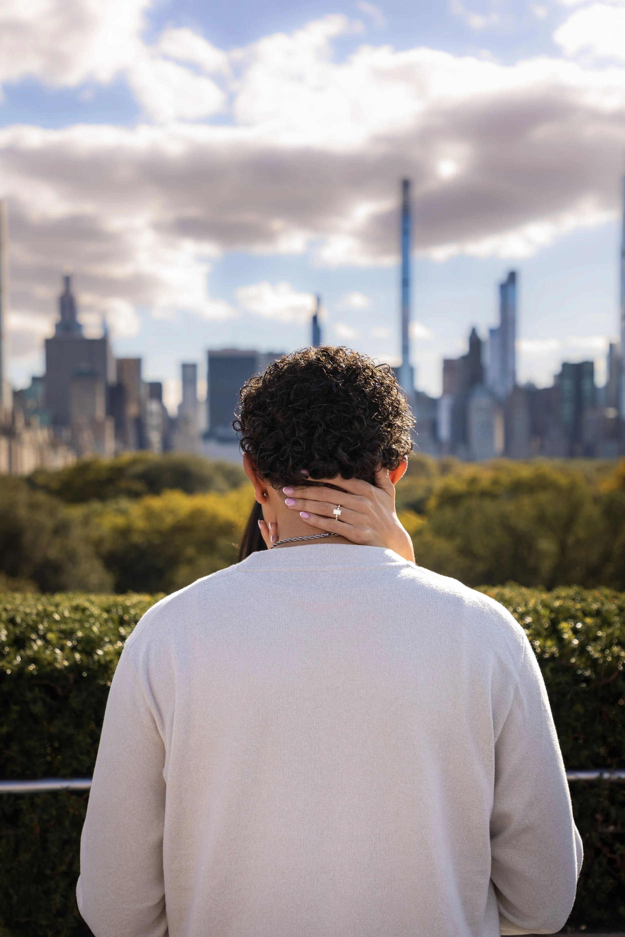 Back view of a person with curly hair holding their head with one hand, in an outdoor setting with a city skyline and partly cloudy sky in the background.
