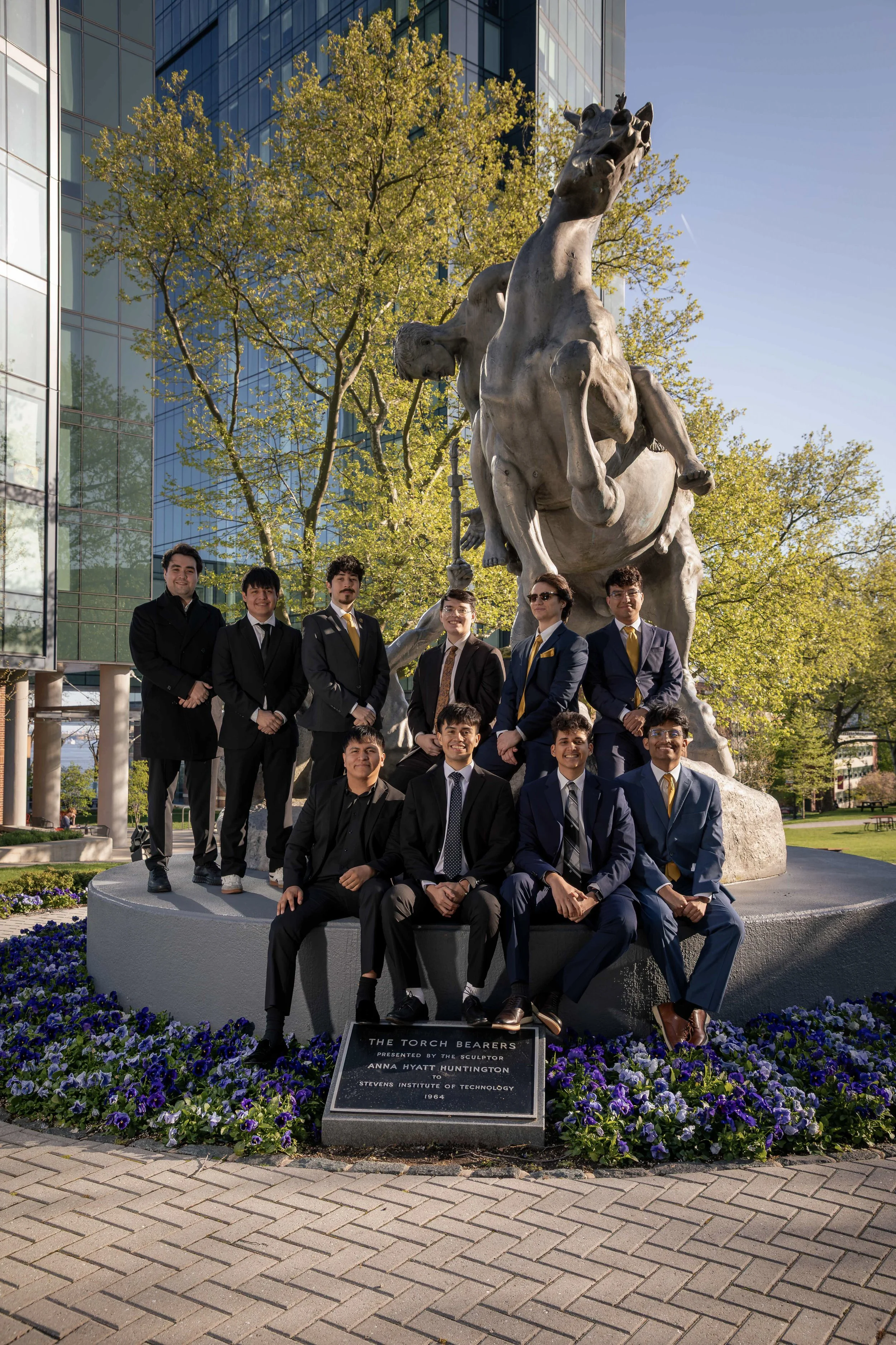 Young men in formal suits gathered around a statue of a horse with a rider, in front of a modern glass building and trees with green leaves, during daytime.