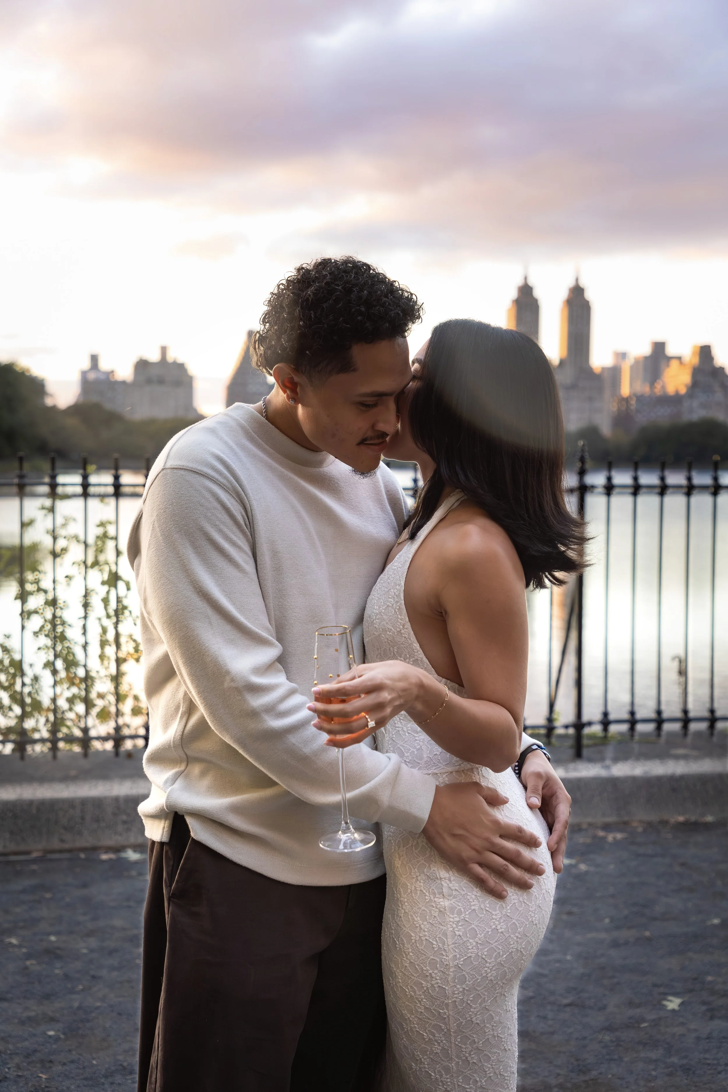 A couple sharing a kiss outdoors during sunset, with city buildings and a river in the background, and the woman holding a glass of champagne.