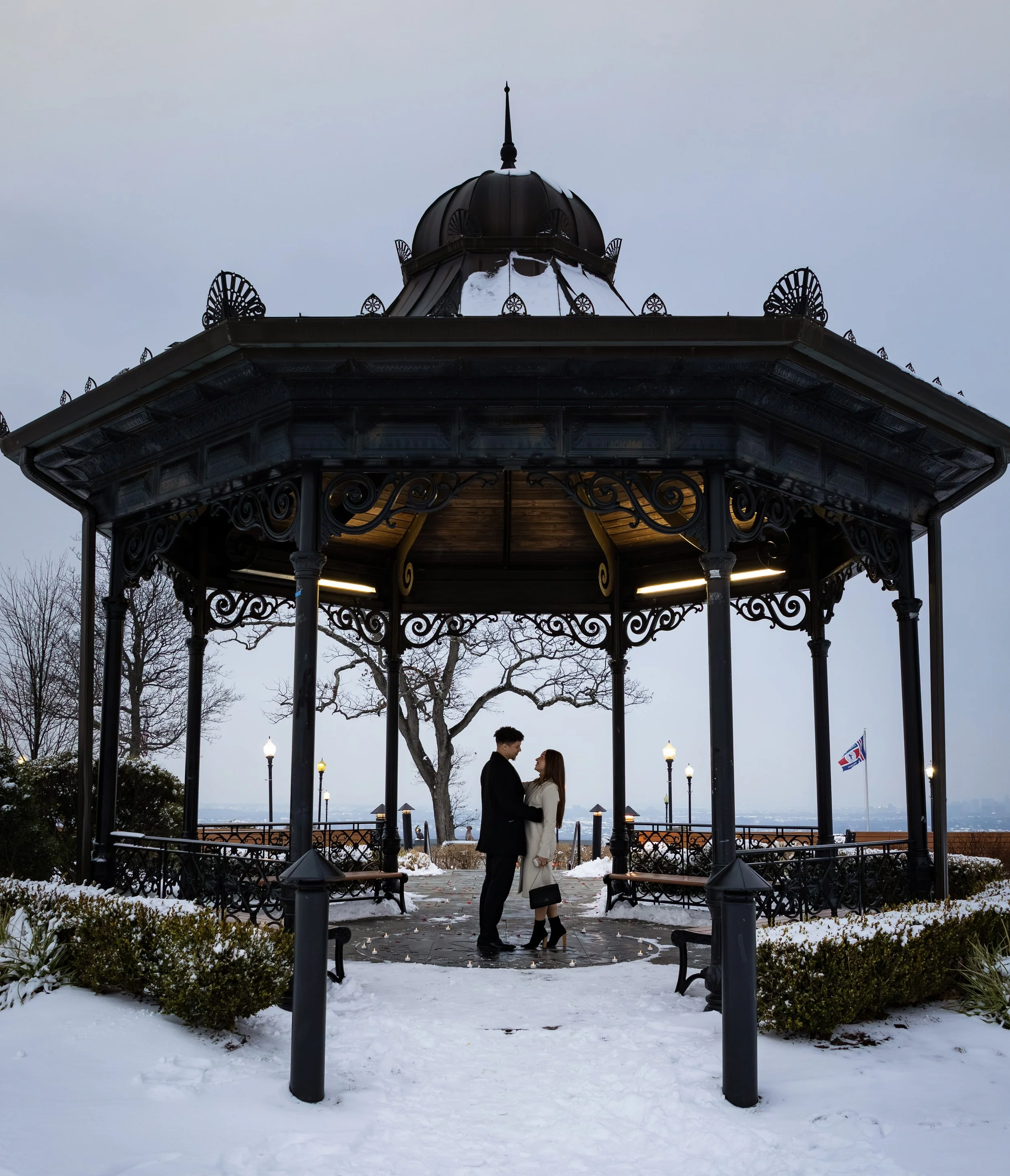A couple stands under a decorative wrought iron gazebo in a snowy park, holding hands and looking into each other's eyes at dusk.