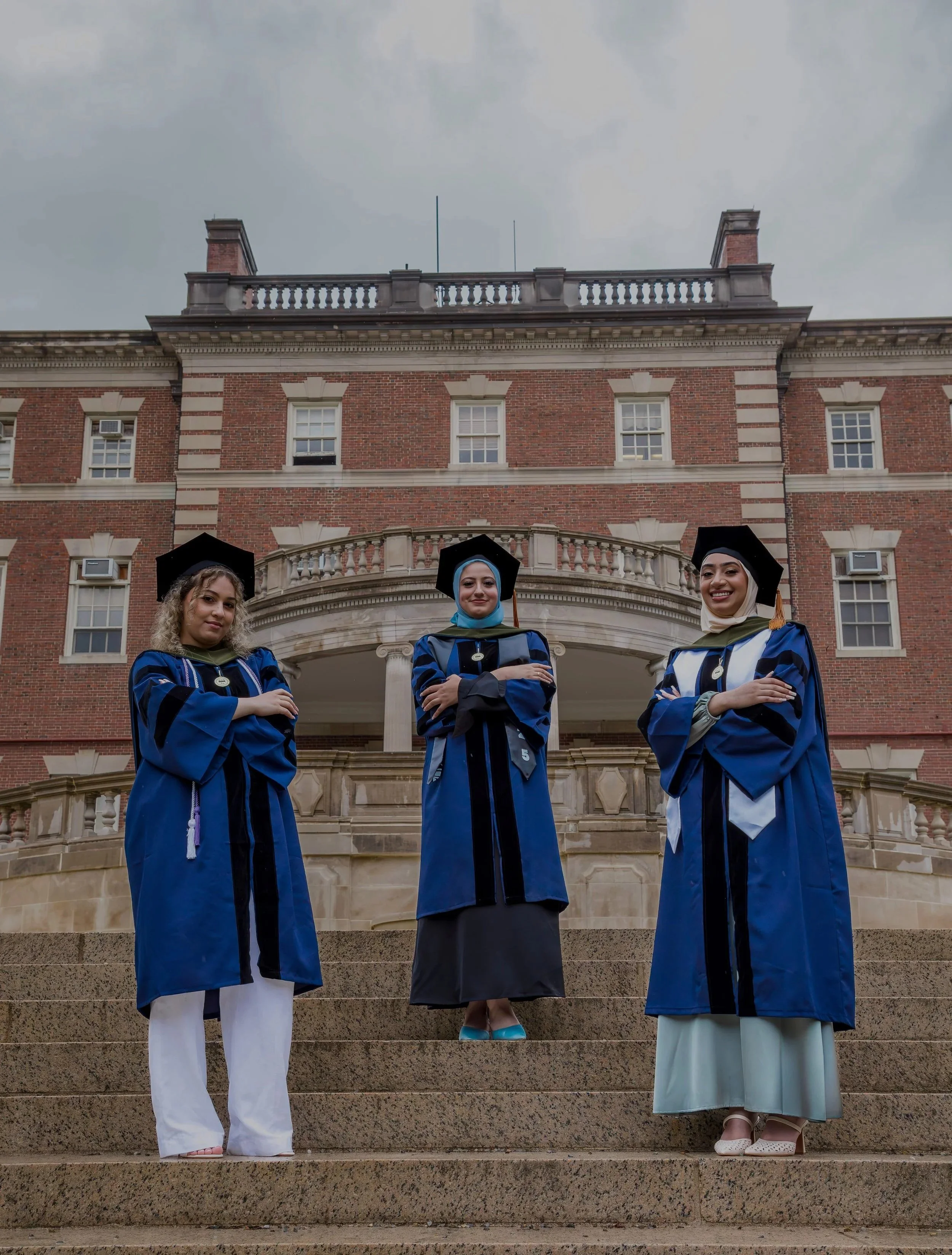 Three women in graduation gowns and caps standing on steps in front of a brick building with a rounded balcony and columns, under cloudy sky.