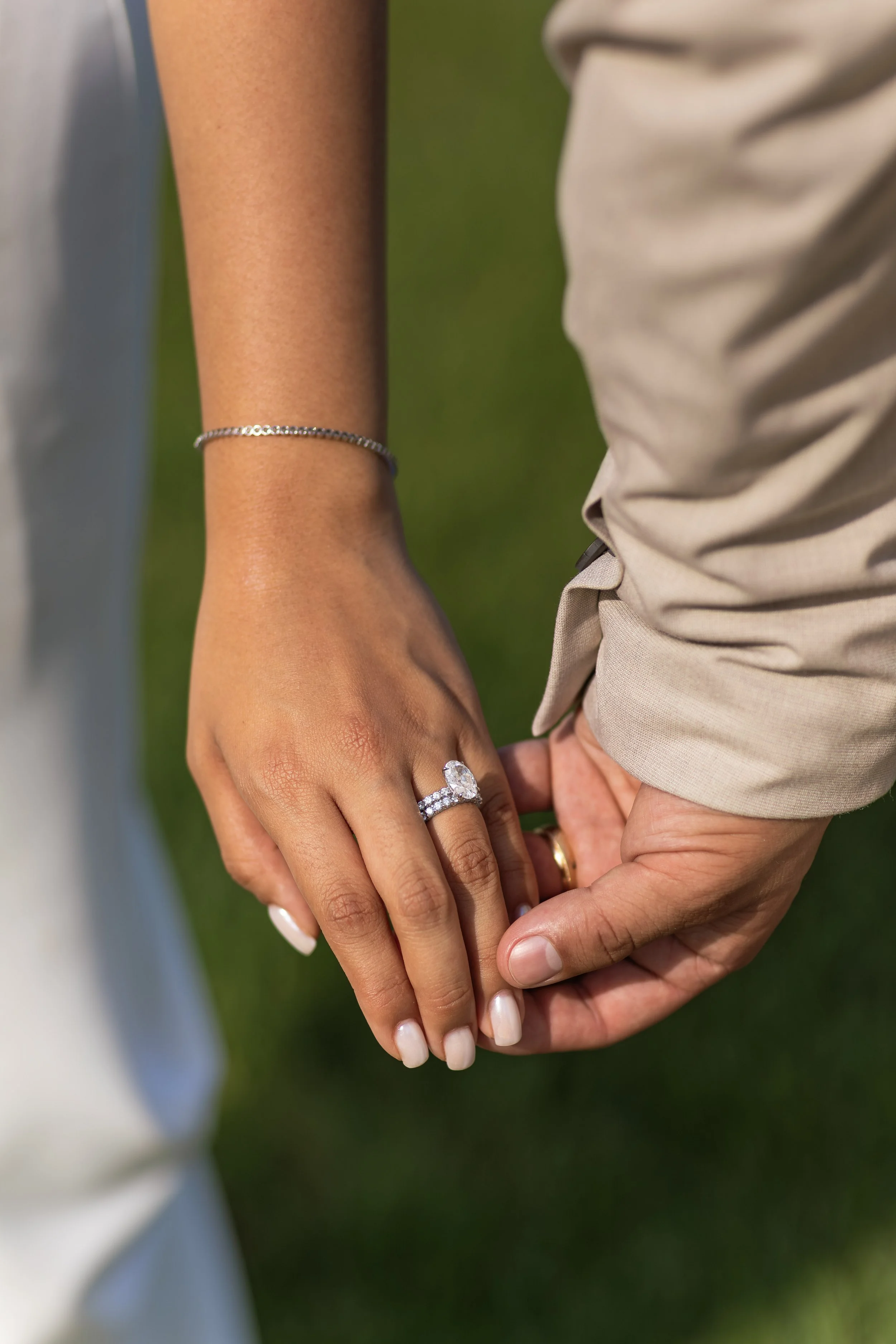 Close-up of a couple holding hands, with the woman's hand showing an engagement ring and a bracelet, against a blurred natural green background.