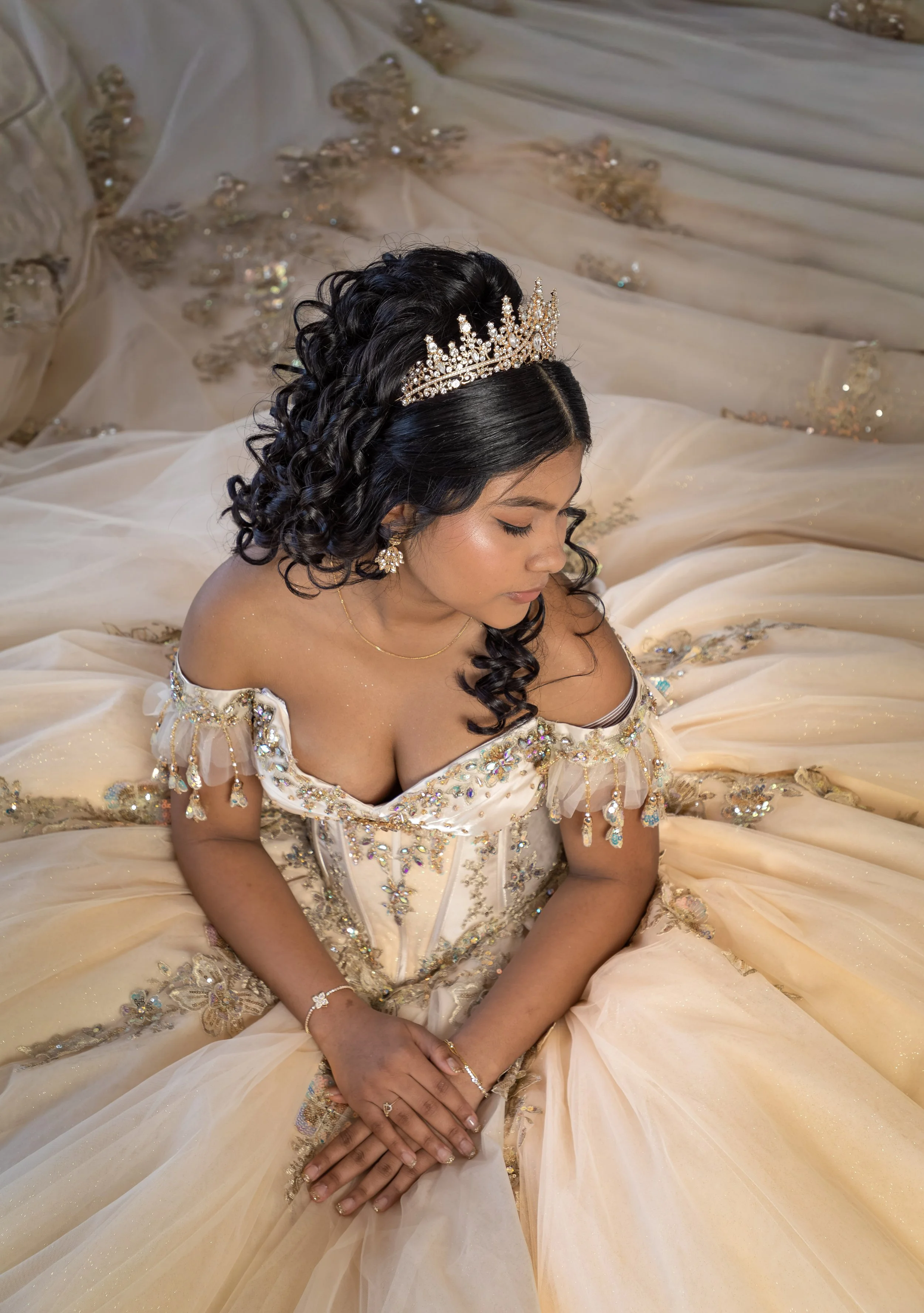 Young woman in an elegant, off-shoulder cream-colored gown decorated with sequins and embroidery, wearing a rhinestone tiara and jewelry, with her eyes closed and her hands resting on her lap, seated on a stage or backdrop with flowing fabric and emb