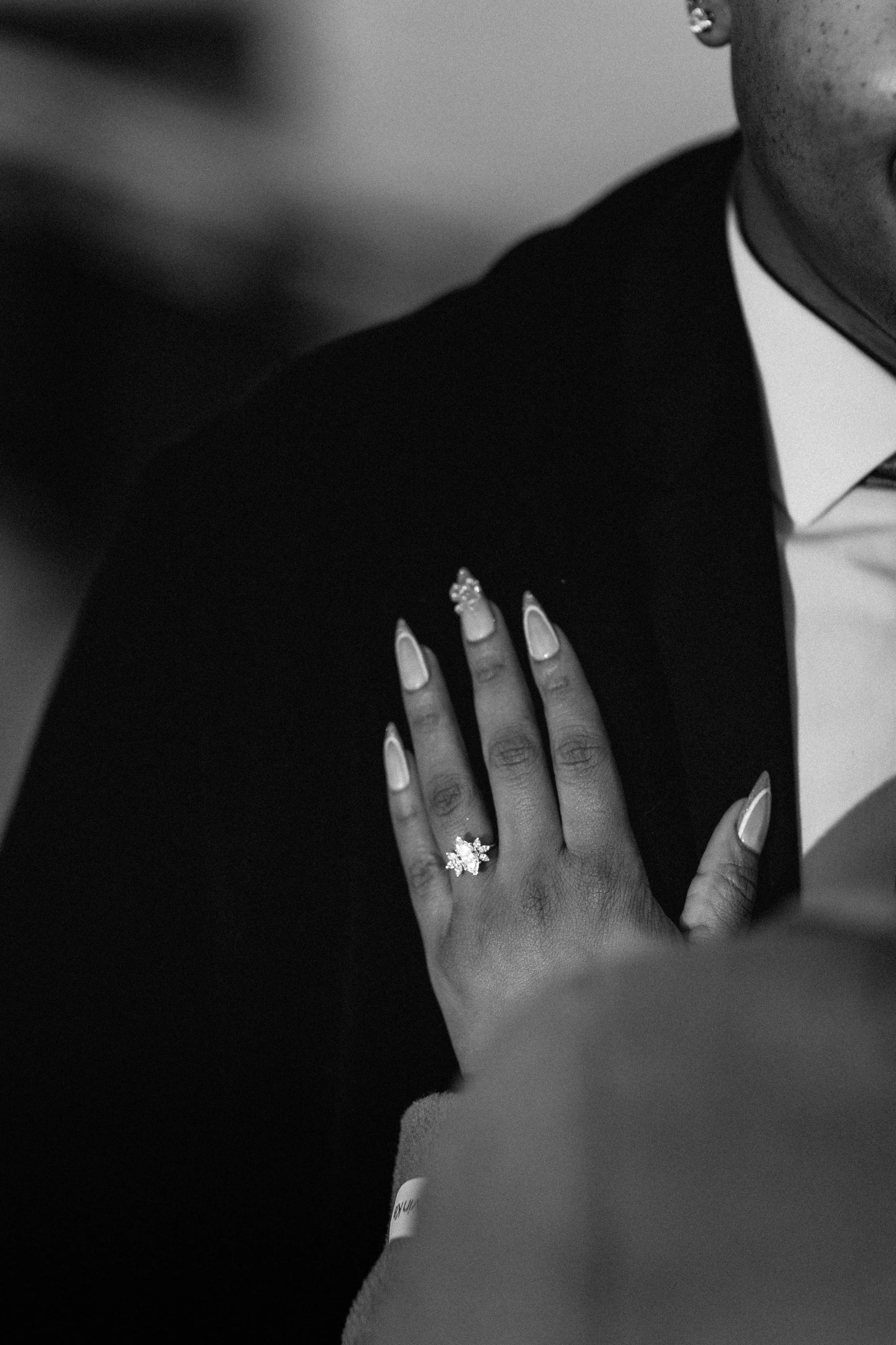 Close-up of a woman's hand with manicured nails and a large diamond engagement ring, resting on a man's chest dressed in a tuxedo, in black and white.