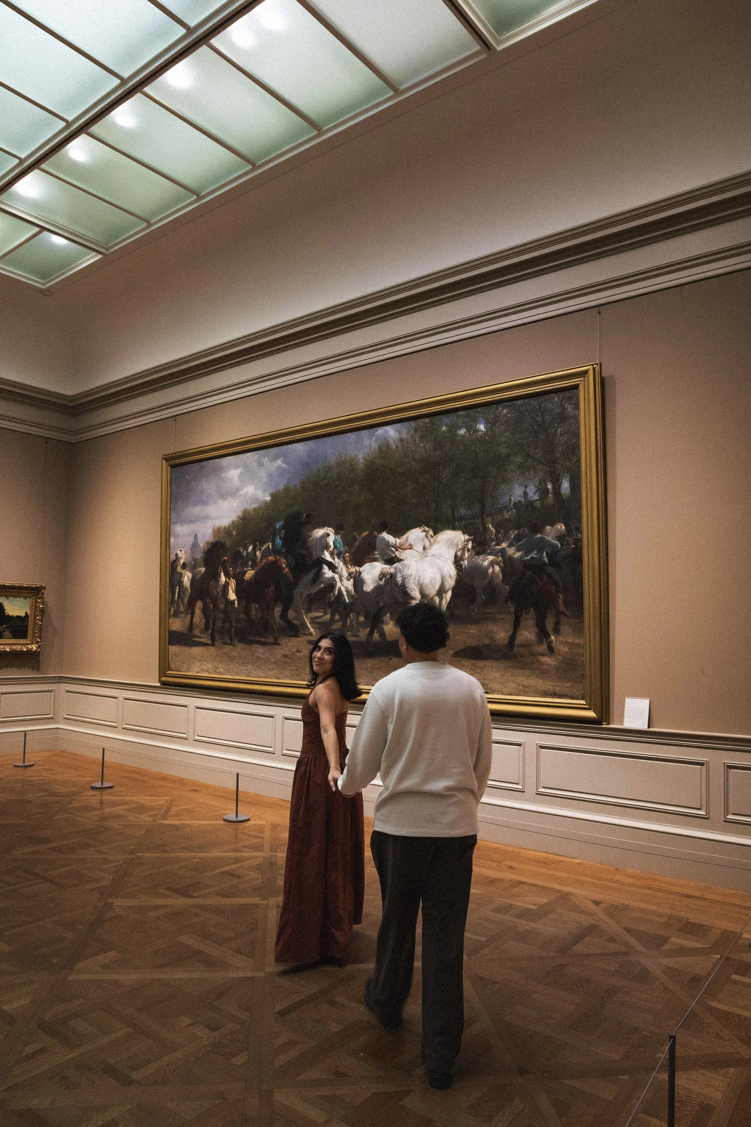 Two people are standing in front of a large painting of a group of horses, with trees and a cloudy sky in the background, inside an art gallery. The woman is wearing a long dress, and the man is wearing a white shirt and dark pants.