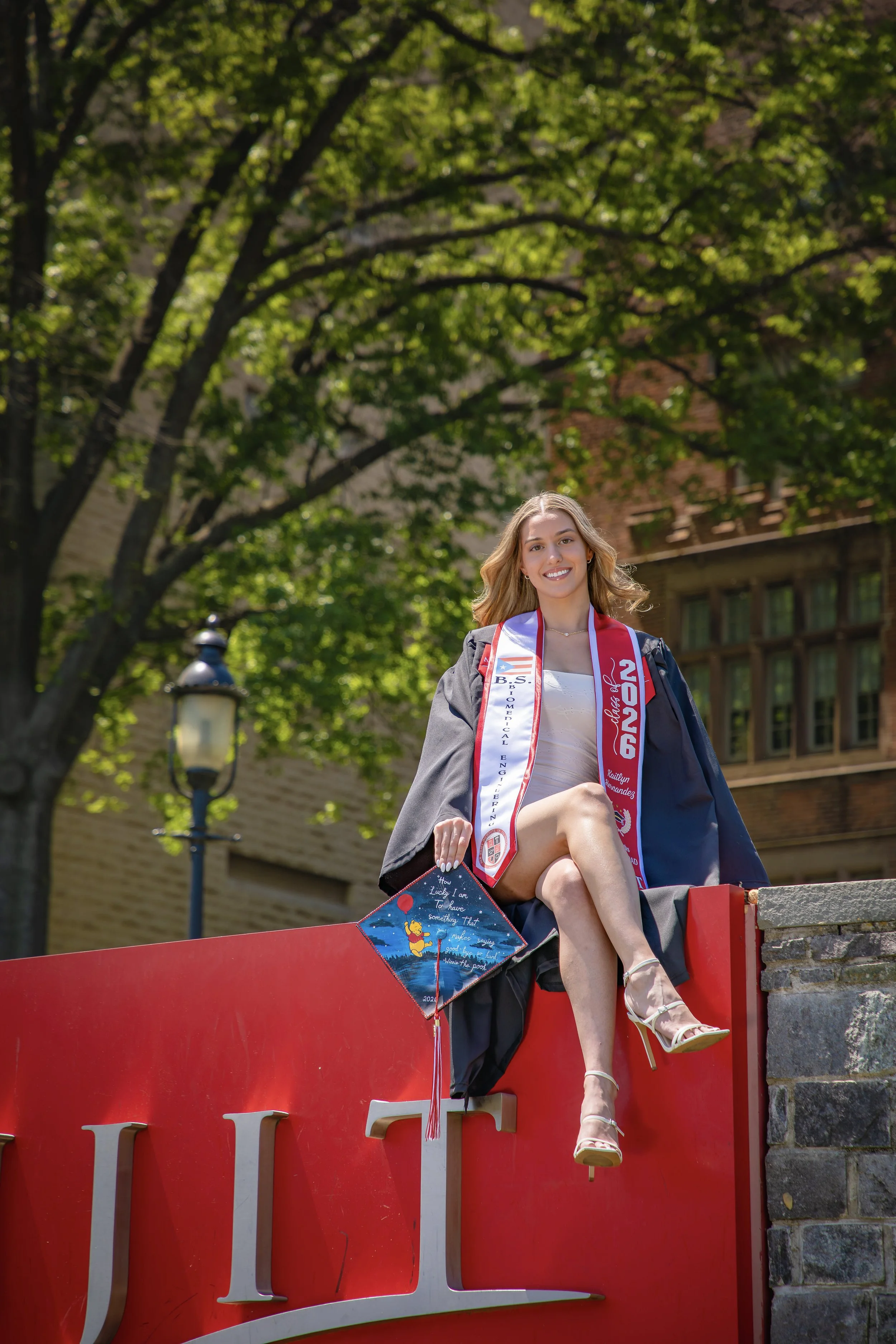 A young woman wearing a graduation gown, cap, and sash sitting on a red ledge outdoors in front of a tree and brick building, smiling at the camera.