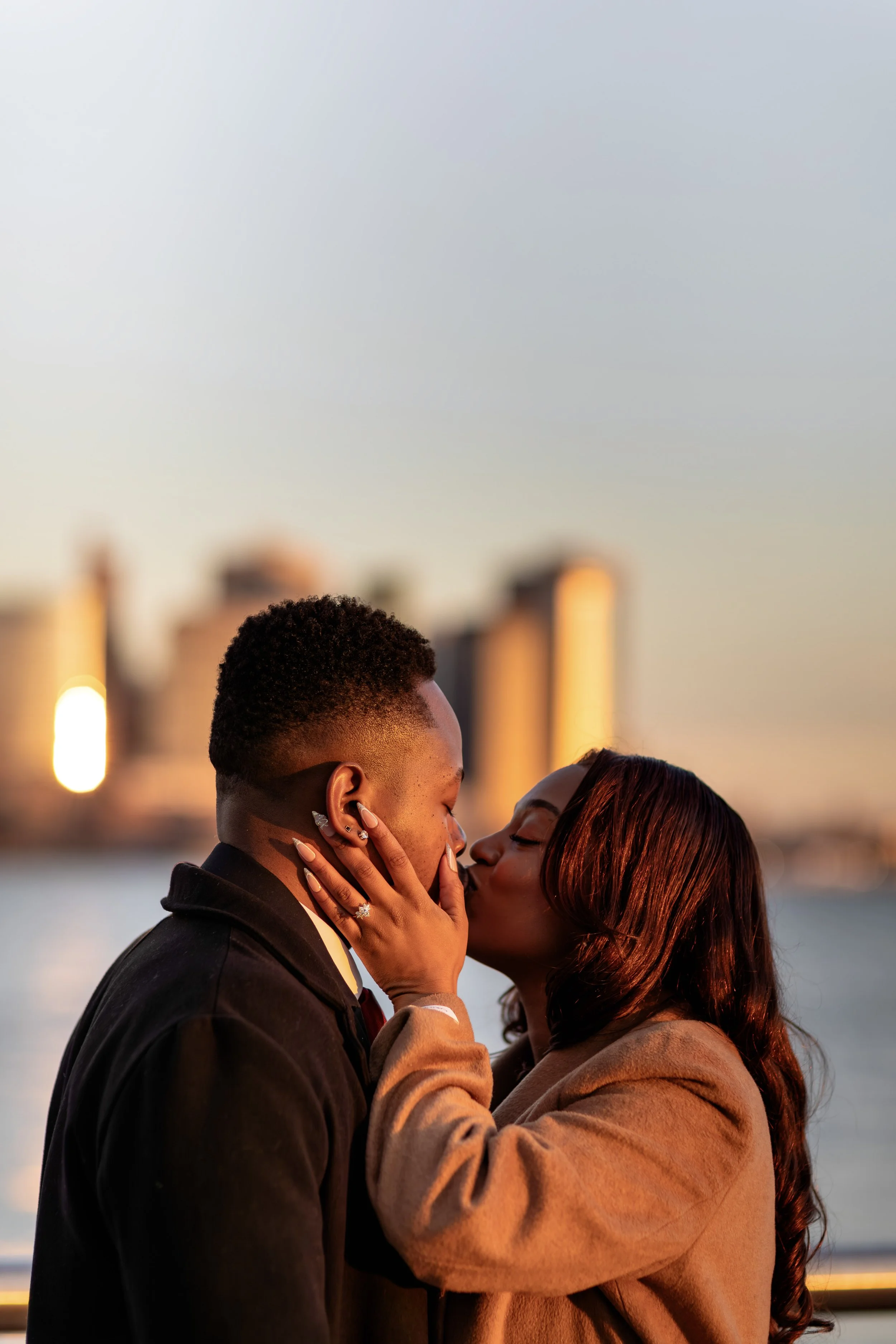 A couple sharing an intimate moment by the water at sunset, facing each other closely and holding each other gently.