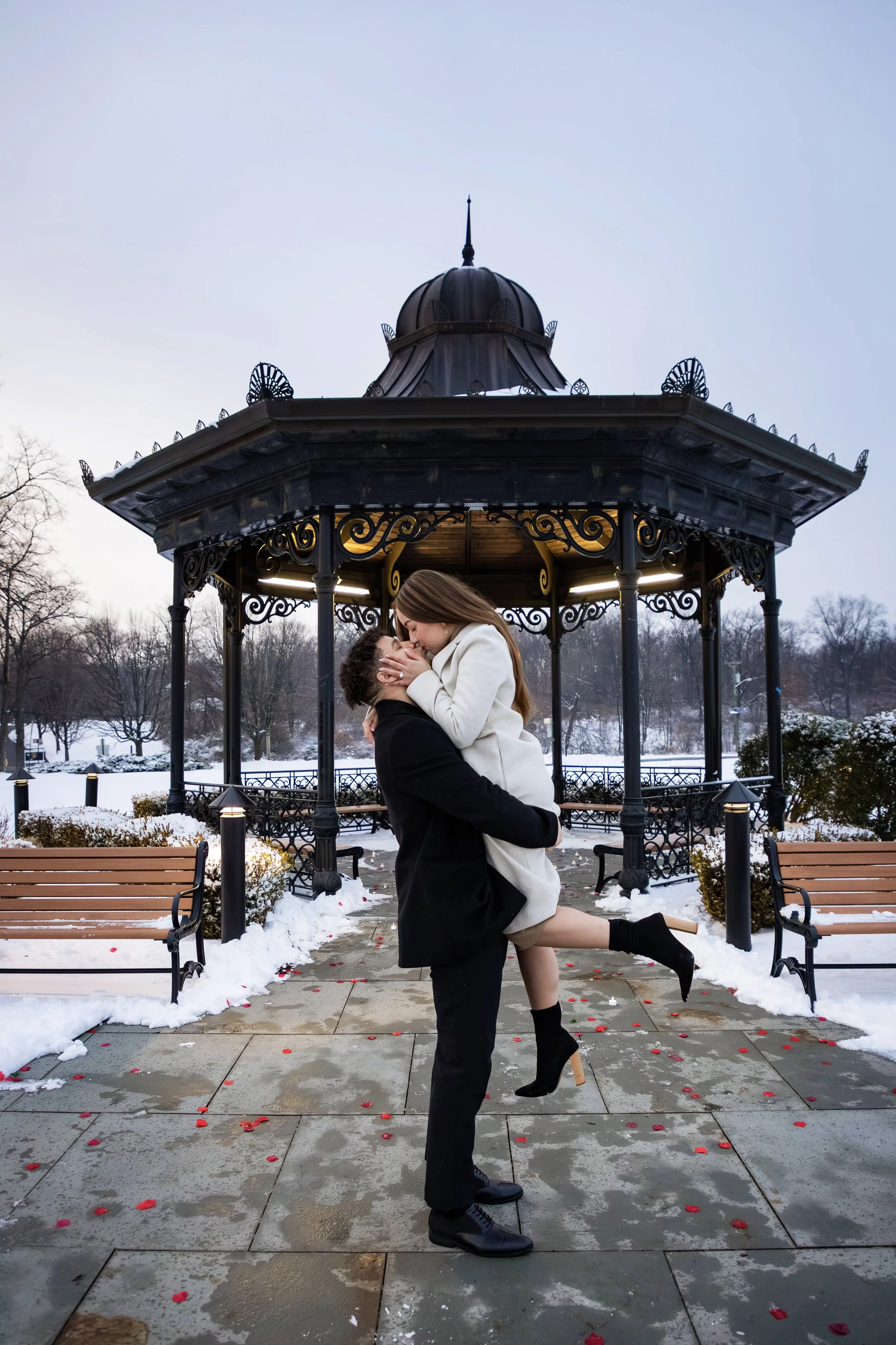 A couple, with the woman in a white coat and the man in a black coat, is sharing a romantic moment outdoors in a snowy park near a decorative pavilion. The woman is being lifted off the ground by the man, and they are embracing and smiling. Rose peta