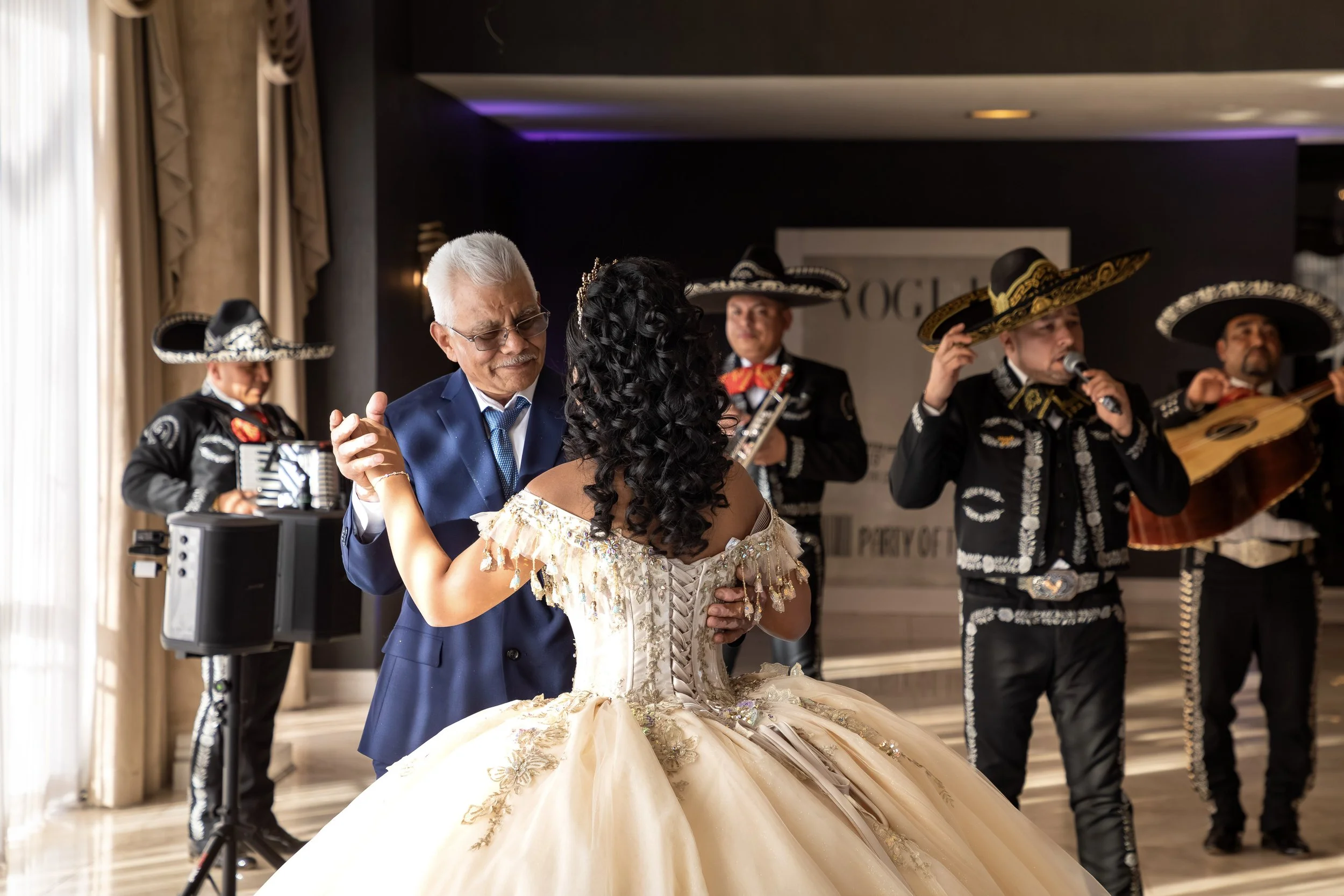 An elderly man in a blue suit and glasses dances with a woman in an elegant, gold gown, while mariachi musicians in traditional black outfits and sombreros perform in the background.