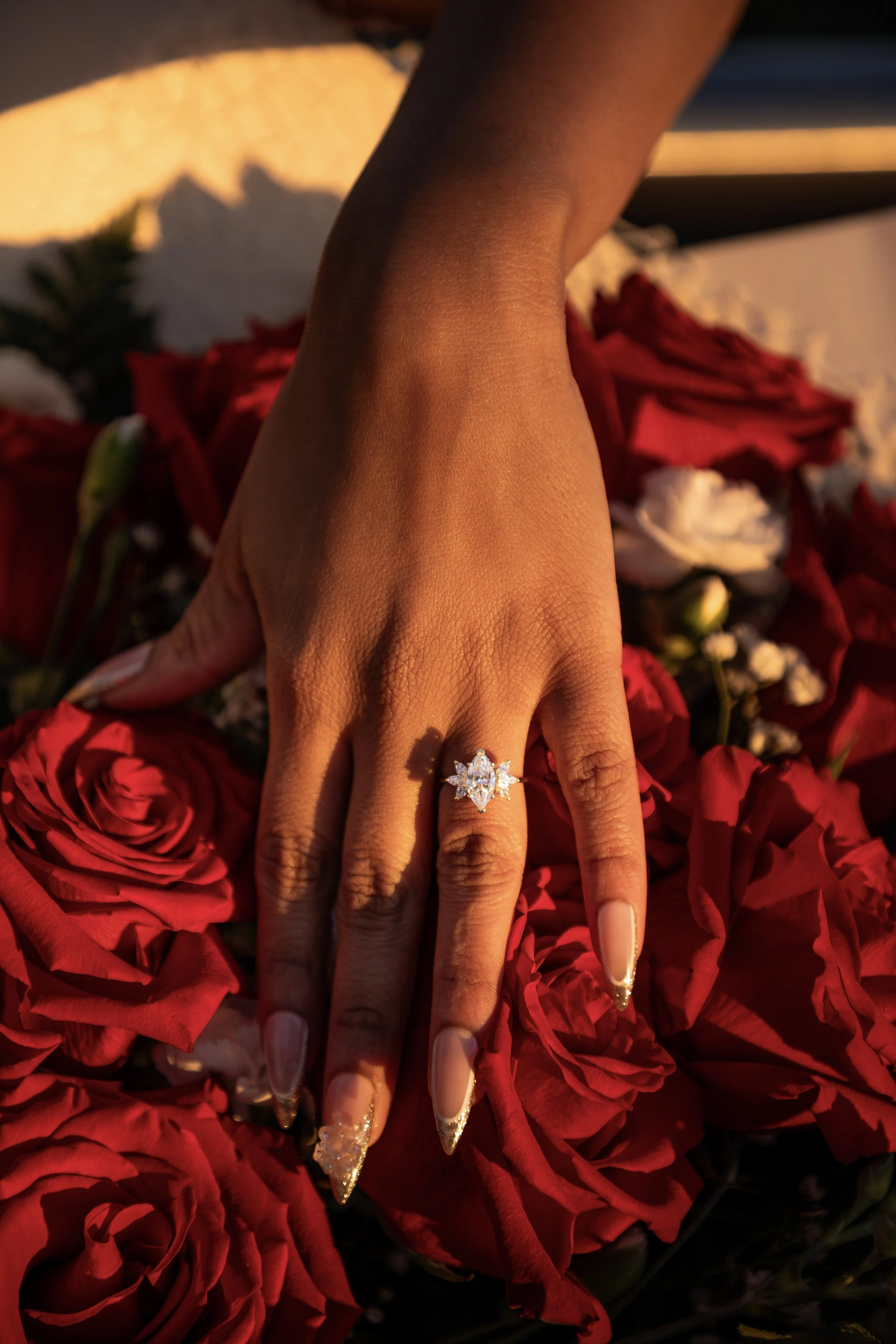 A woman's hand with a large, ornate diamond ring on her ring finger, resting on red roses. The photo is taken in warm sunlight, highlighting her nails and skin tone.