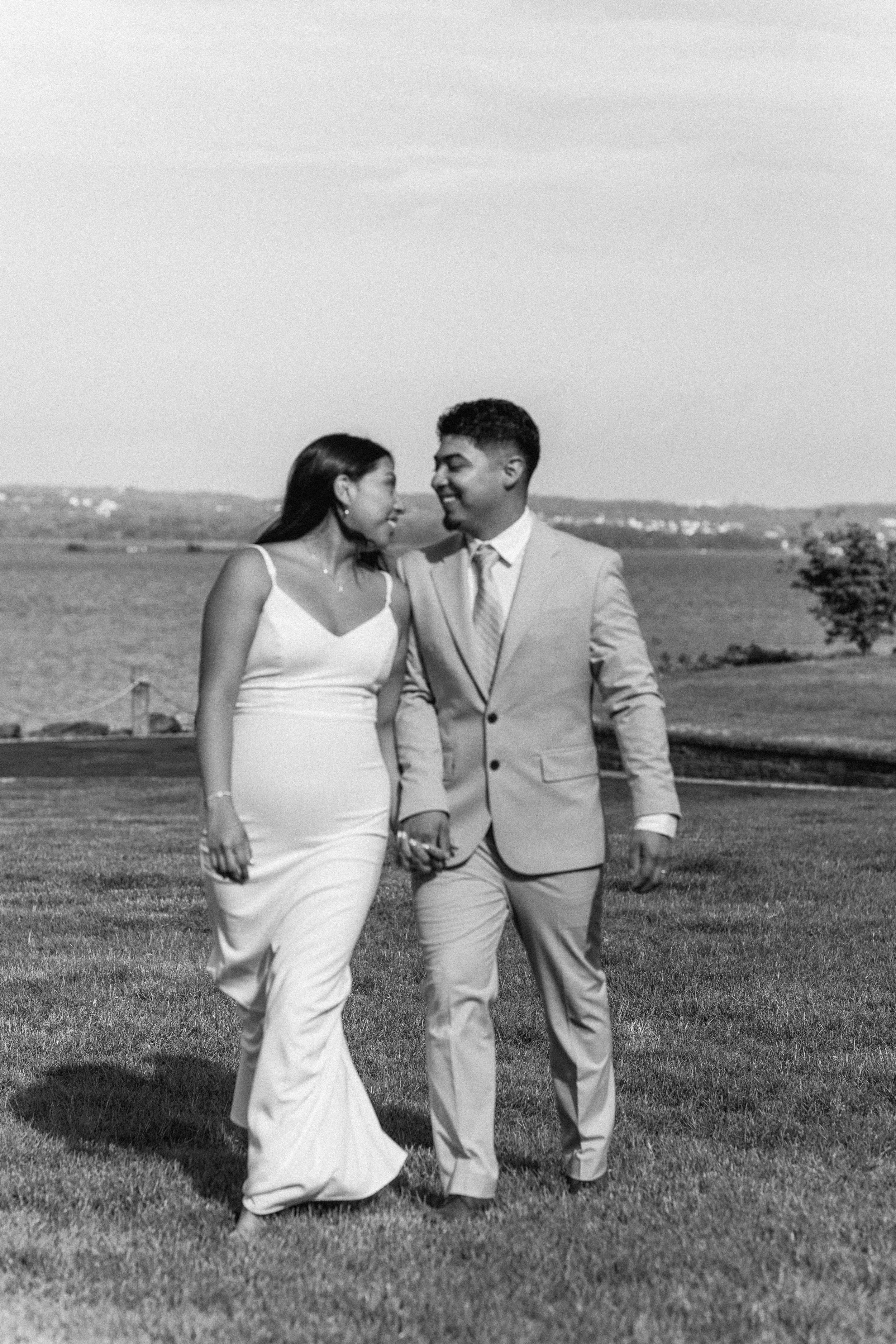 A black and white photo of a couple walking hand in hand outdoors, smiling at each other, with a body of water and hills in the background.