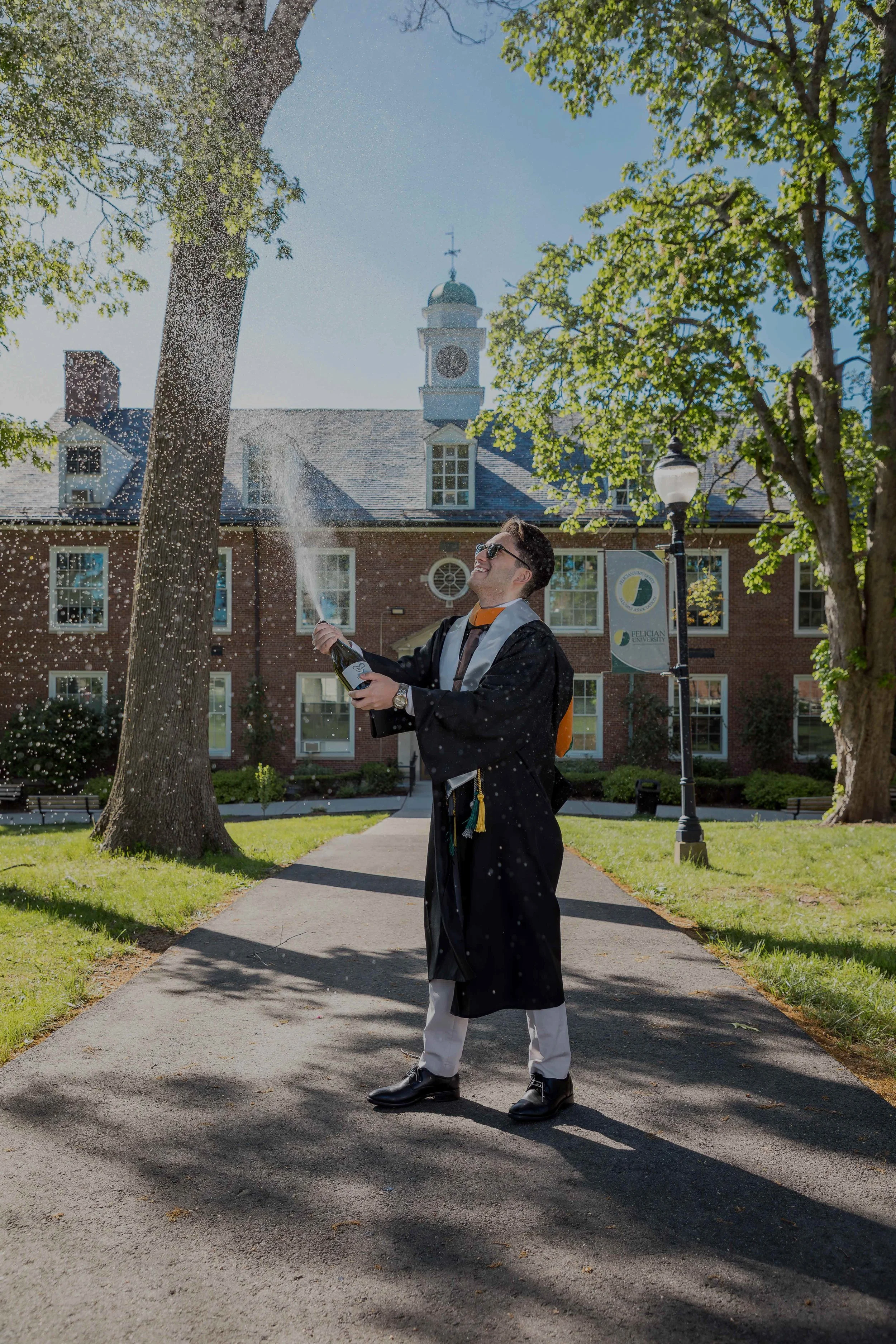 A man in graduation attire celebrating on campus, spraying champagne. Buildings and trees are visible in the background.