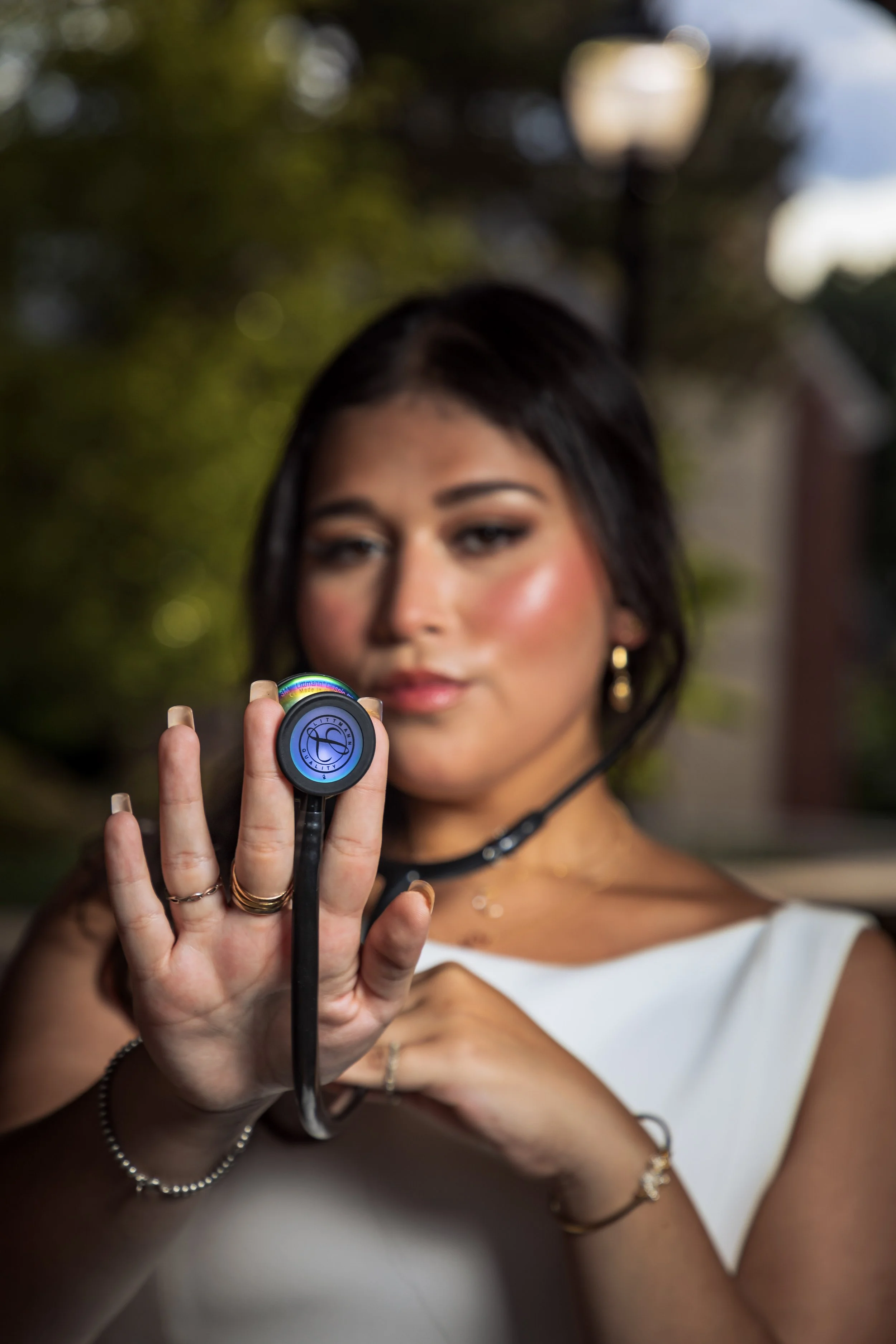 A woman with dark hair, gold hoop earrings, and jewelry on her hands and wrist, is holding a stethoscope toward the camera.