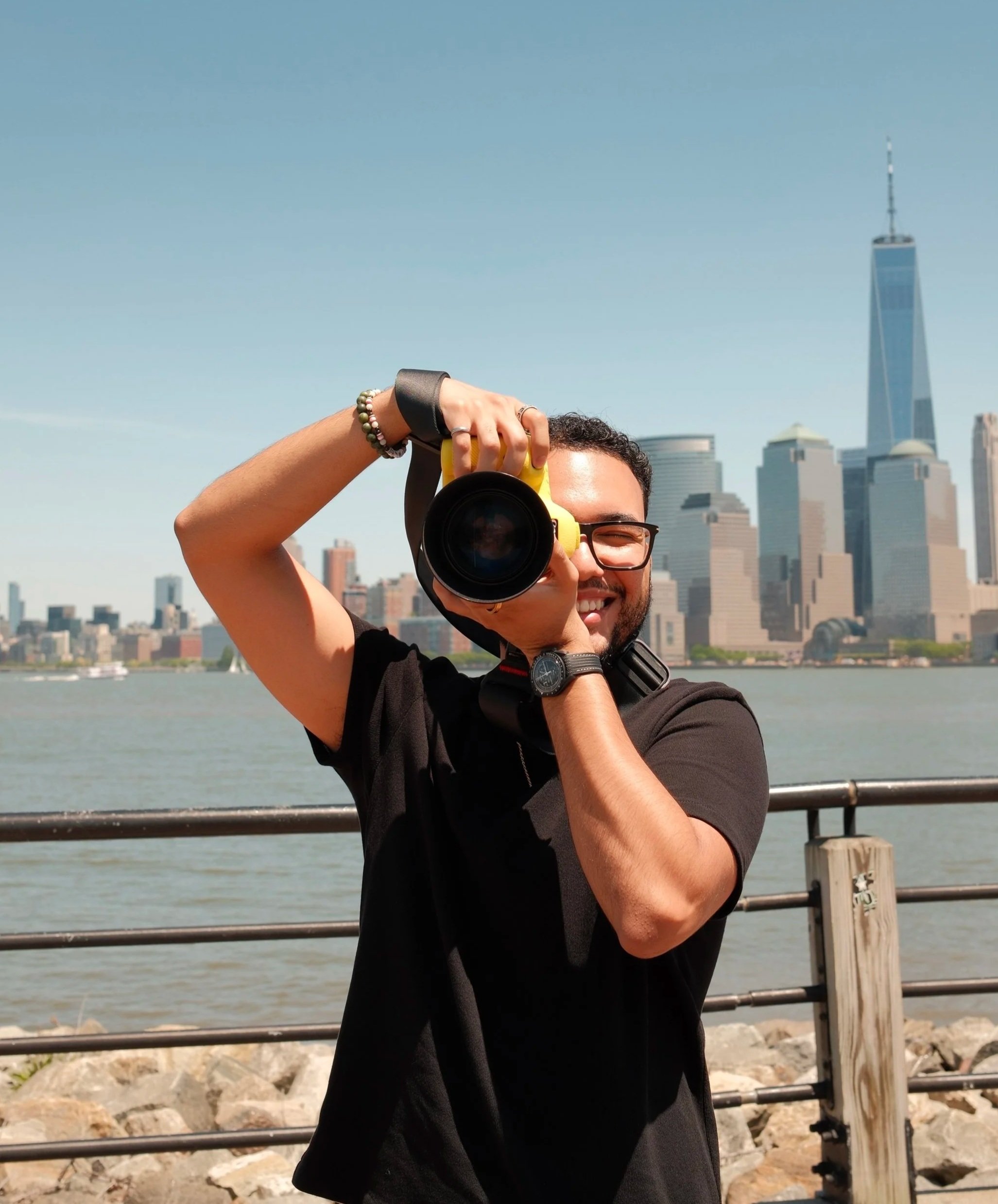 A person taking a photo with a camera on a waterfront with the New York City skyline, including One World Trade Center, in the background.