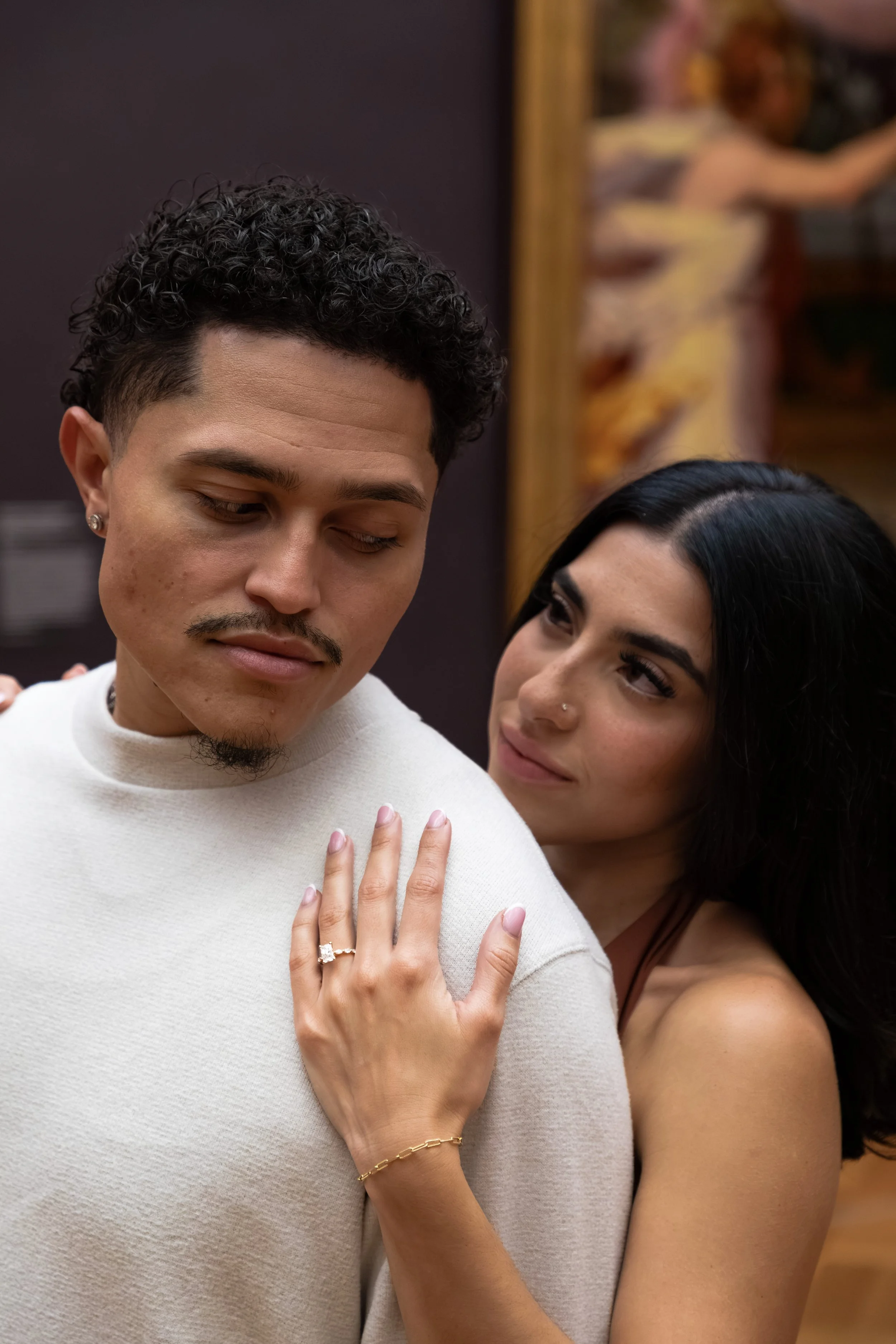 A man with curly dark hair and a mustache shows a woman his left hand with an engagement ring while she embraces him from behind. The woman has black hair, a nose piercing, and is smiling at the man. The woman is wearing a thin strap top, and the man