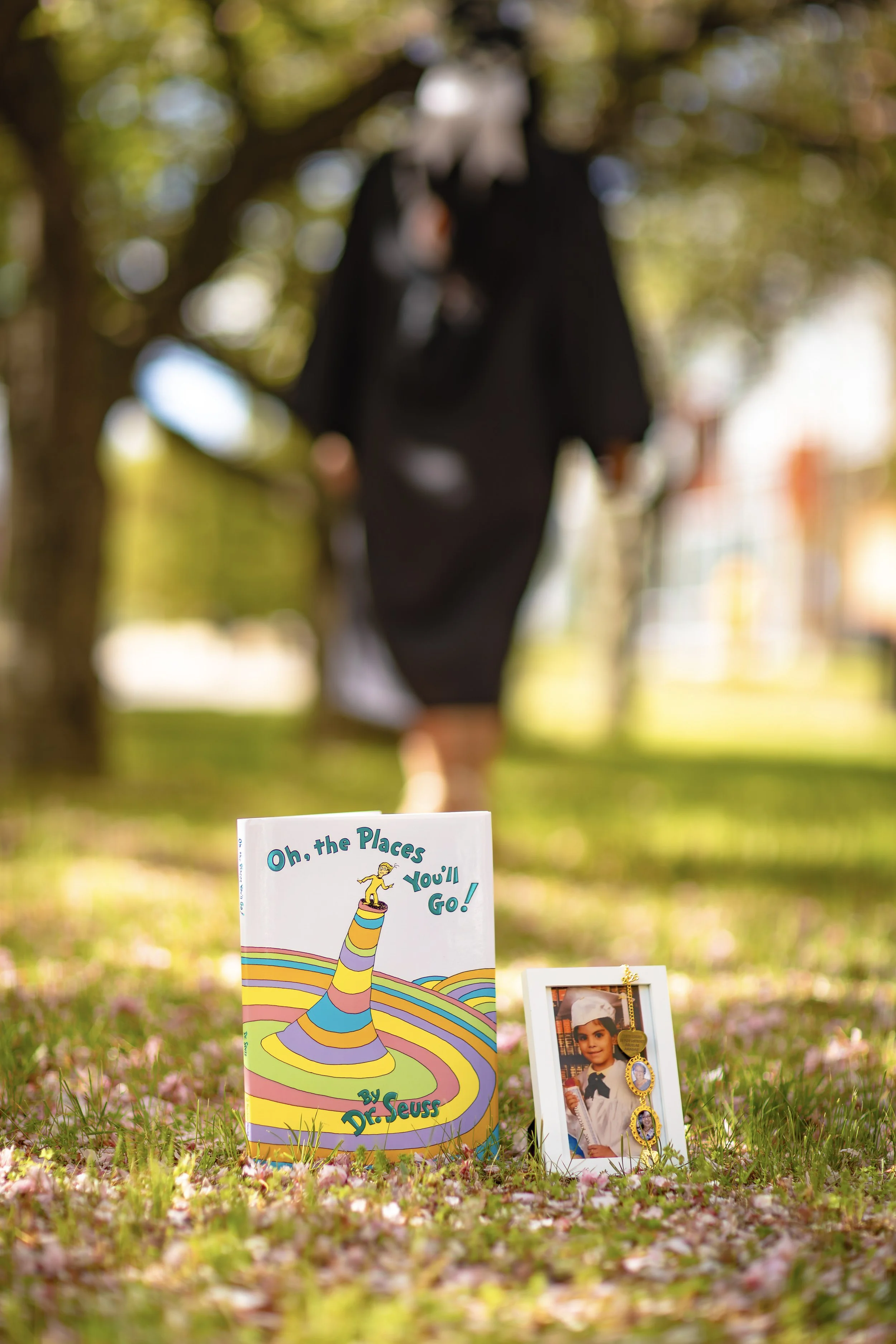 A colorful children's book titled "Oh, the Places You'll Go!" by Dr. Seuss is displayed on grassy ground with pink flower petals, with a framed photograph and medals nearby. In the background, a person dressed in black with a gray hood is blurred, st