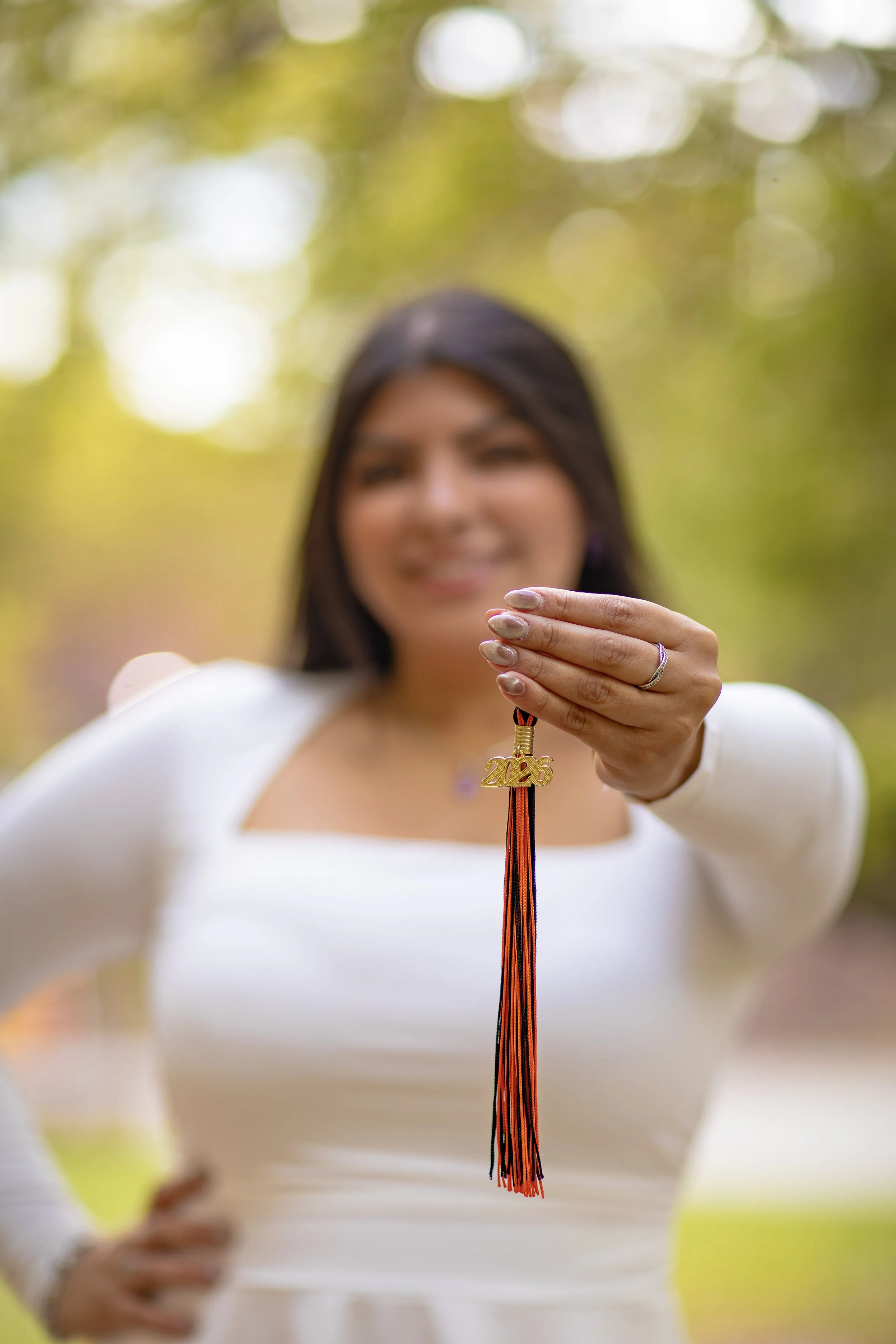 Woman holding a graduation cap with the year 2026 in gold pendant hanging from a black and orange tassel, blurred background with sunlight filtering through trees.