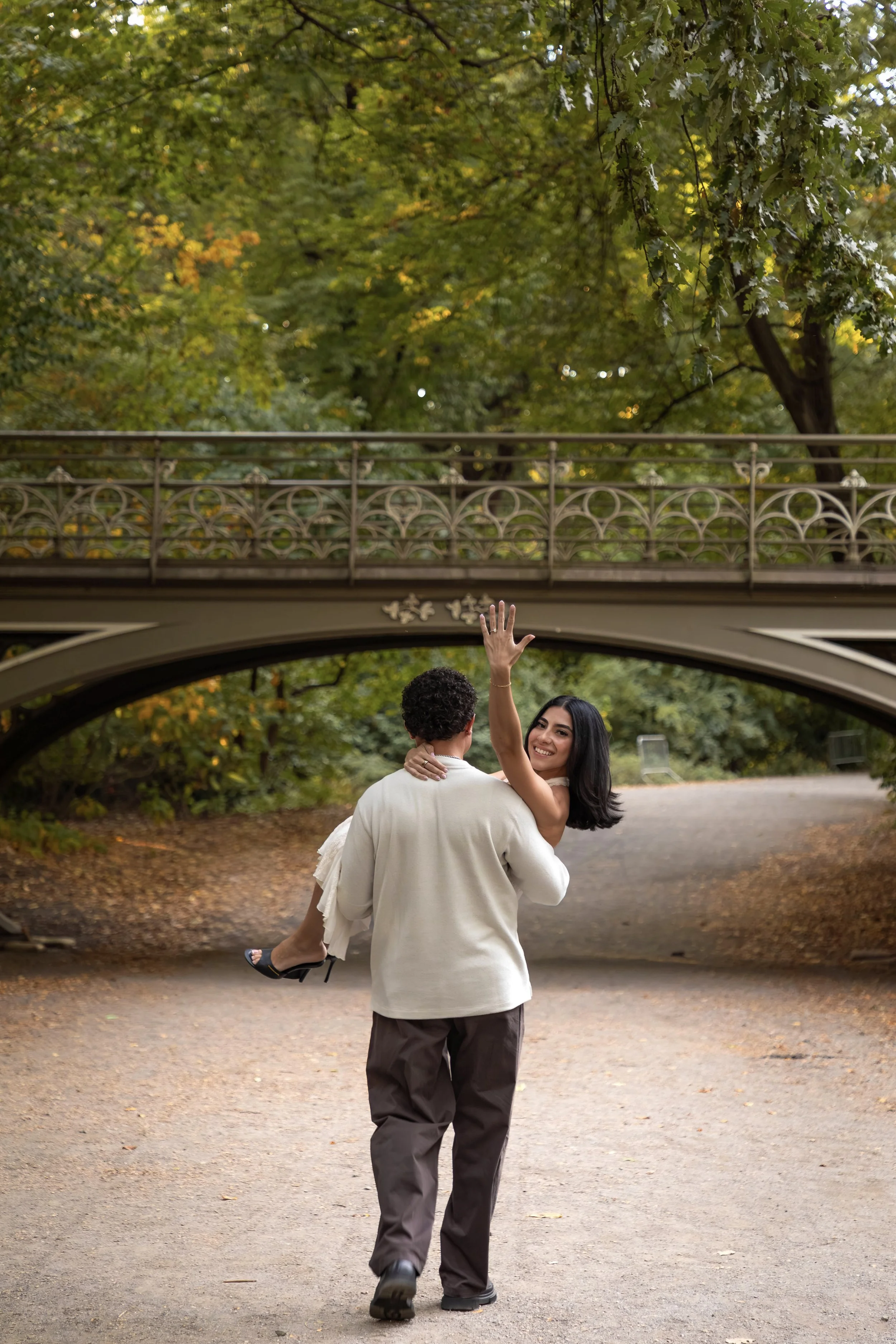 A man is walking away while carrying a smiling woman in a white dress. The woman is raising her hand, and they are walking under a bridge in a park with trees and falling leaves.