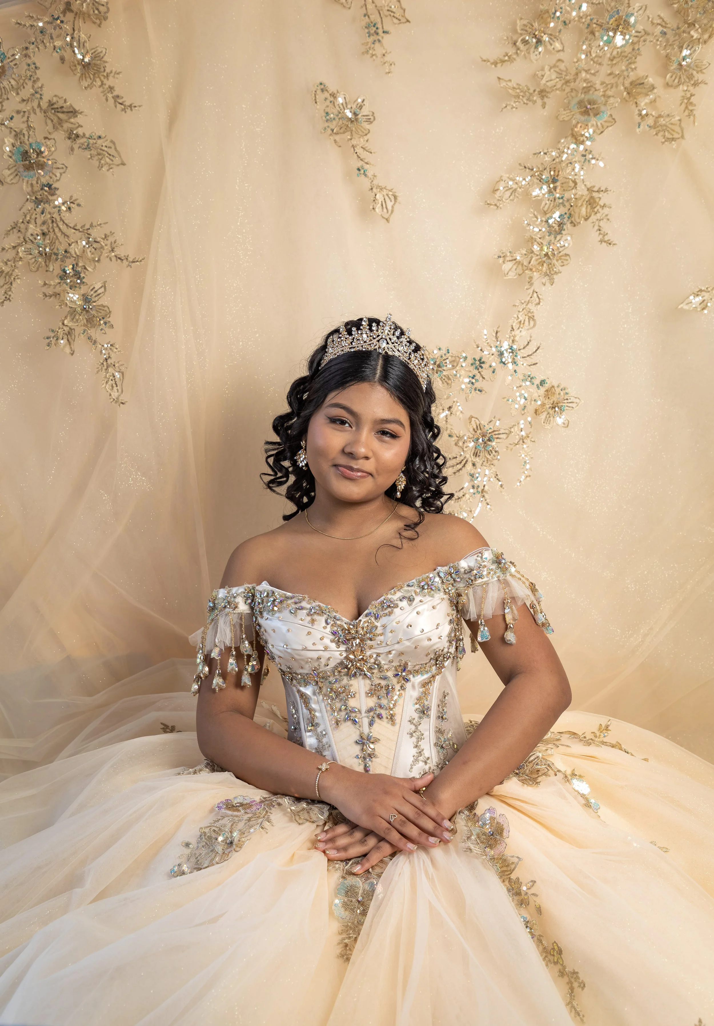 A young woman in a cream-colored, embellished gown with intricate beadwork, lace, and rhinestones, sitting in front of a matching ornate backdrop with gold and blue embroidery and butterfly motifs. She wears a tiara, earrings, and a delicate bracelet