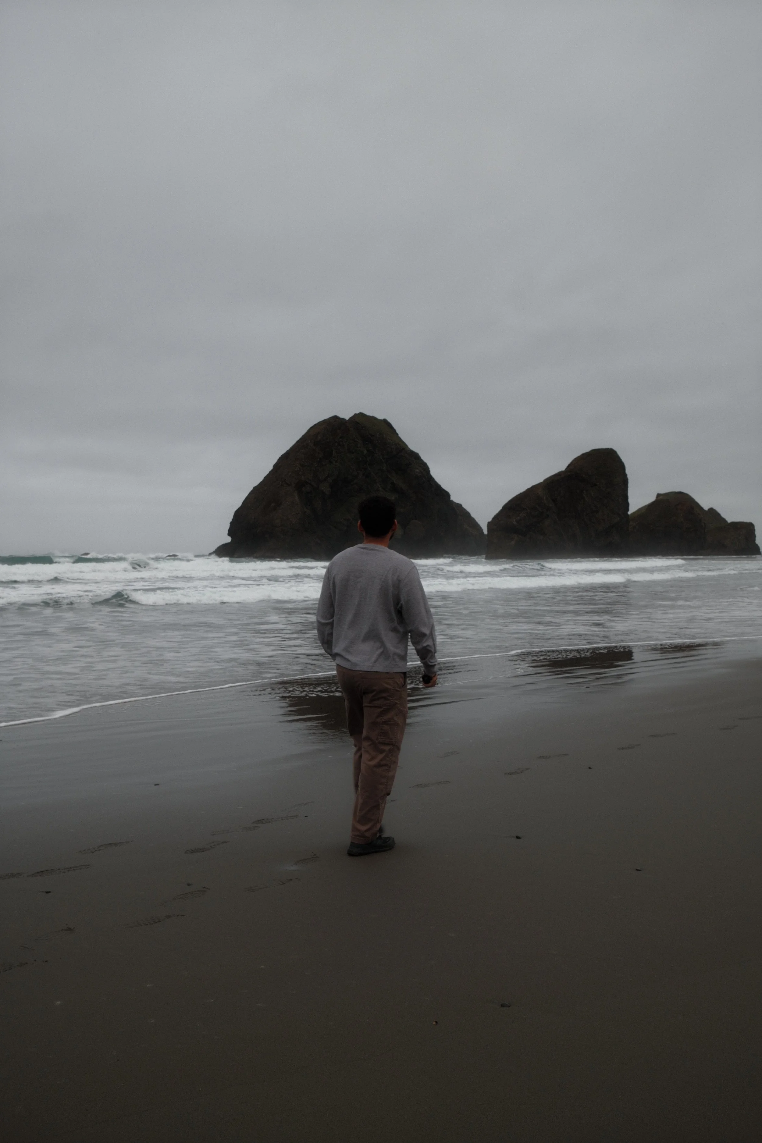 A person standing on a dark sandy beach facing the ocean with large rocks in the background and cloudy gray sky.