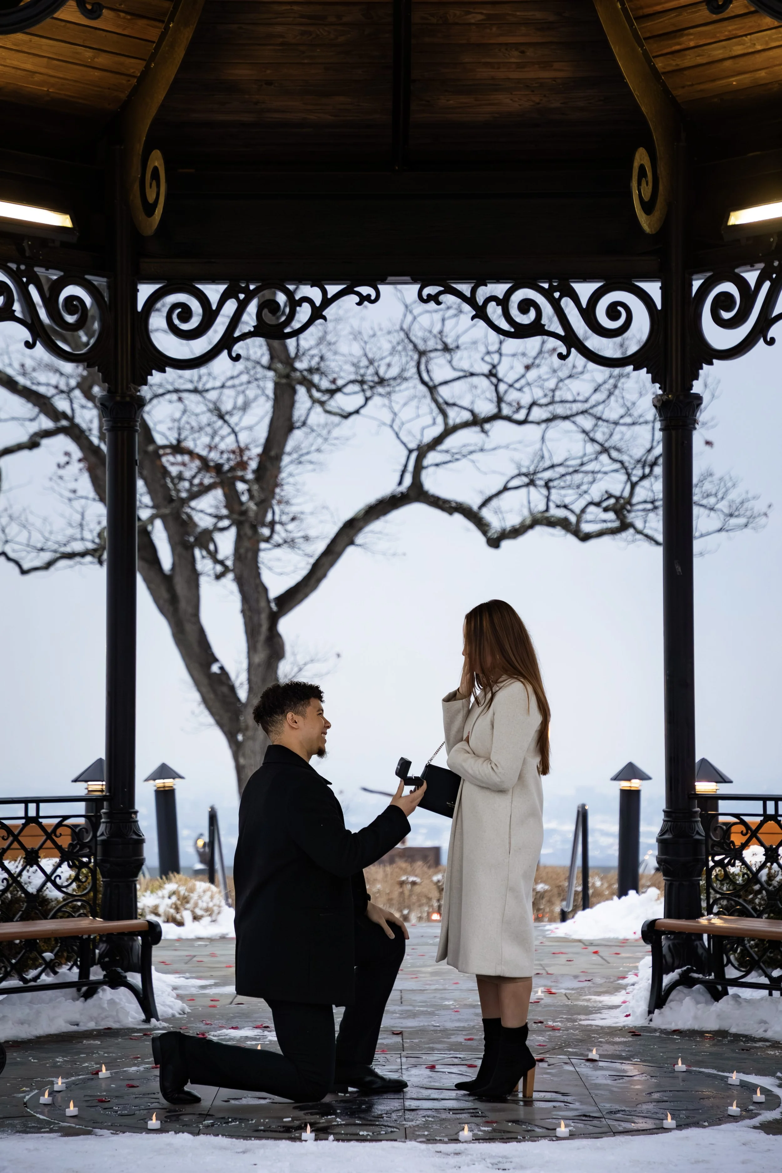 A man on one knee proposing marriage to a woman surrounded by candles on the ground, with a man presenting a ring and a woman reactively covering her face with her hands in an outdoor, snowy setting during evening.