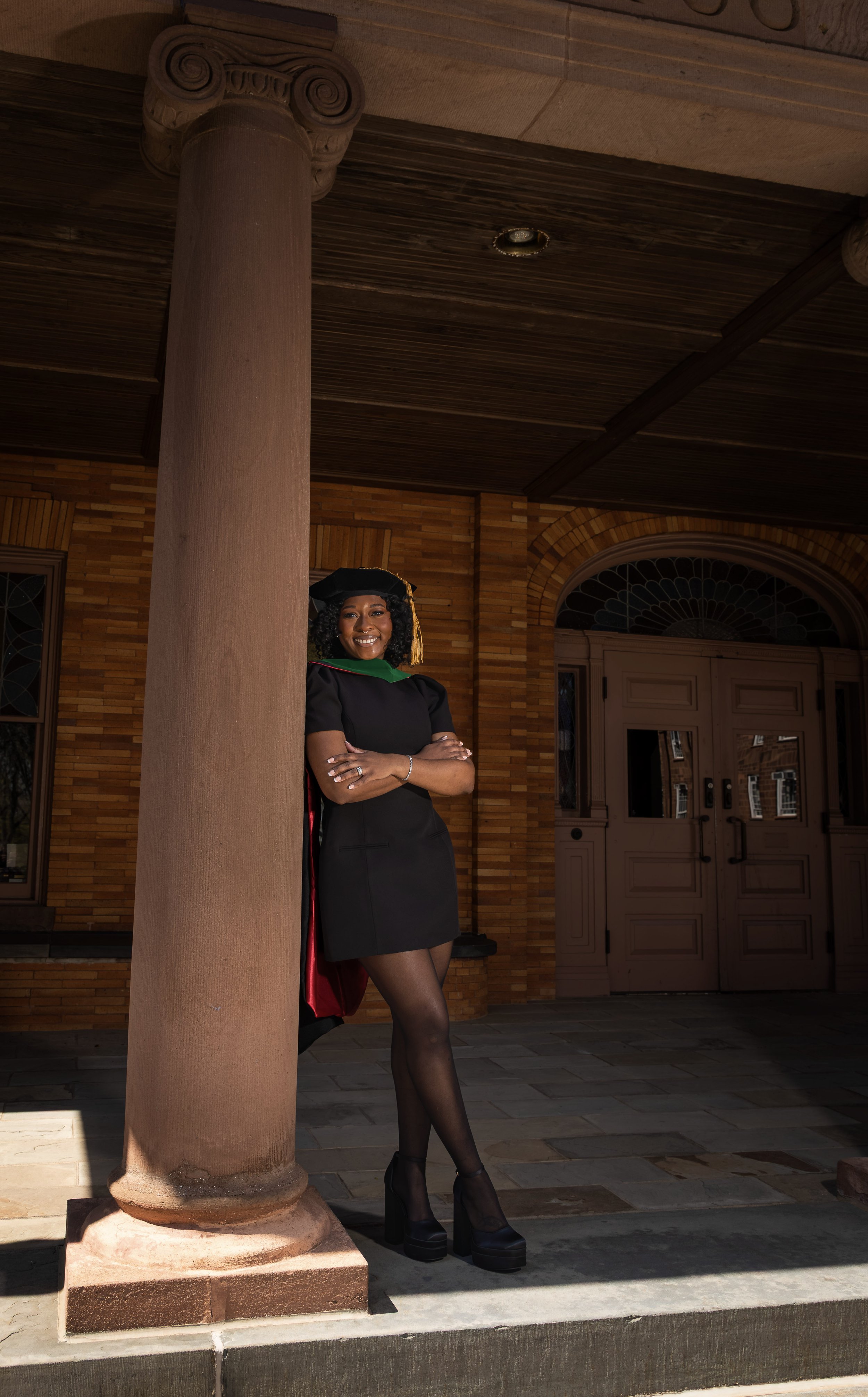 A woman in a graduation cap and gown standing outdoors near a brick building with arched windows.